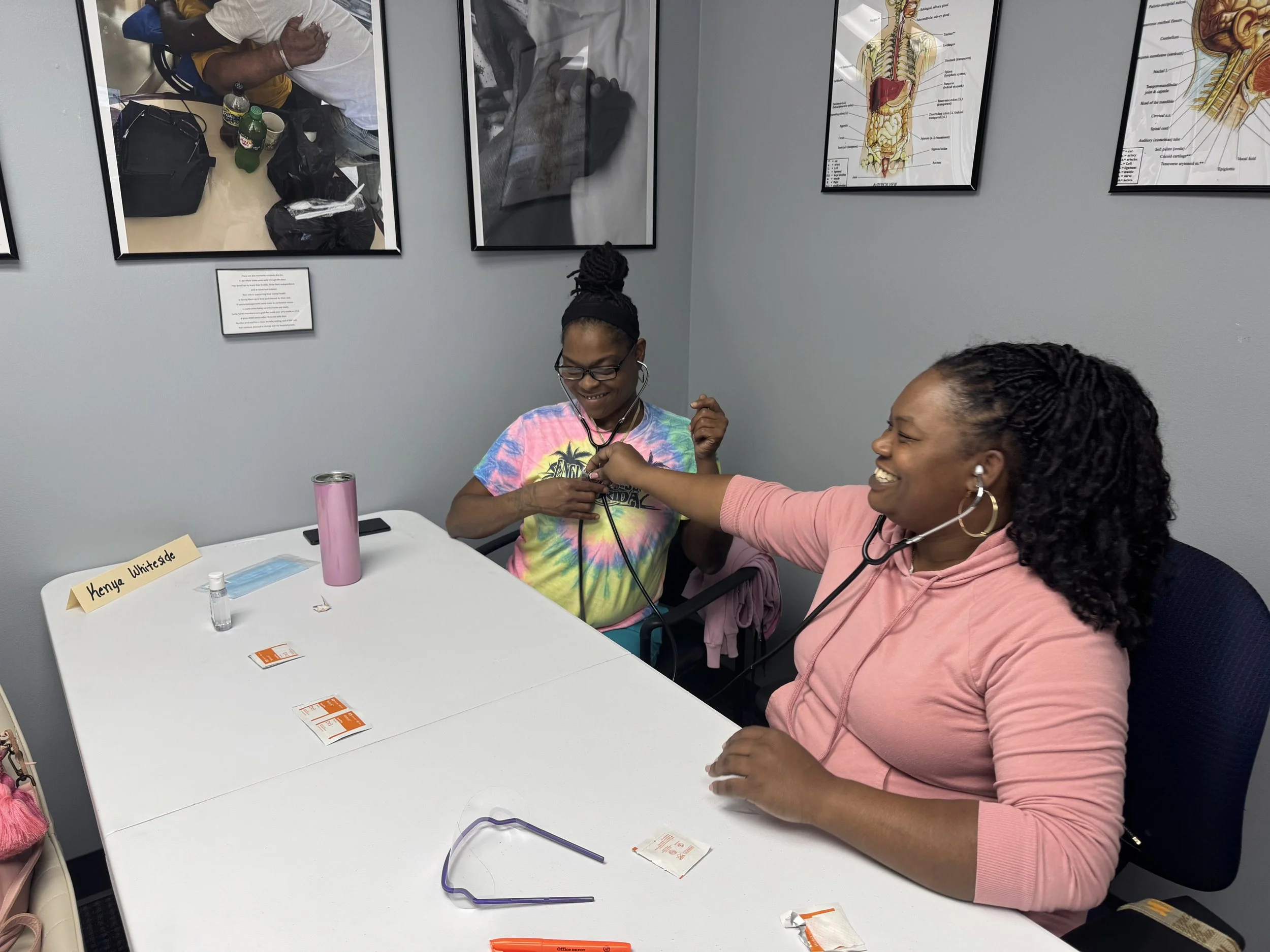 Two women smiling and engaging in conversation at a table in a clinical or educational setting. One woman is wearing a tie-dye shirt, and the other is in a pink hoodie. There are medical items on the table, and framed anatomical posters are hanging o