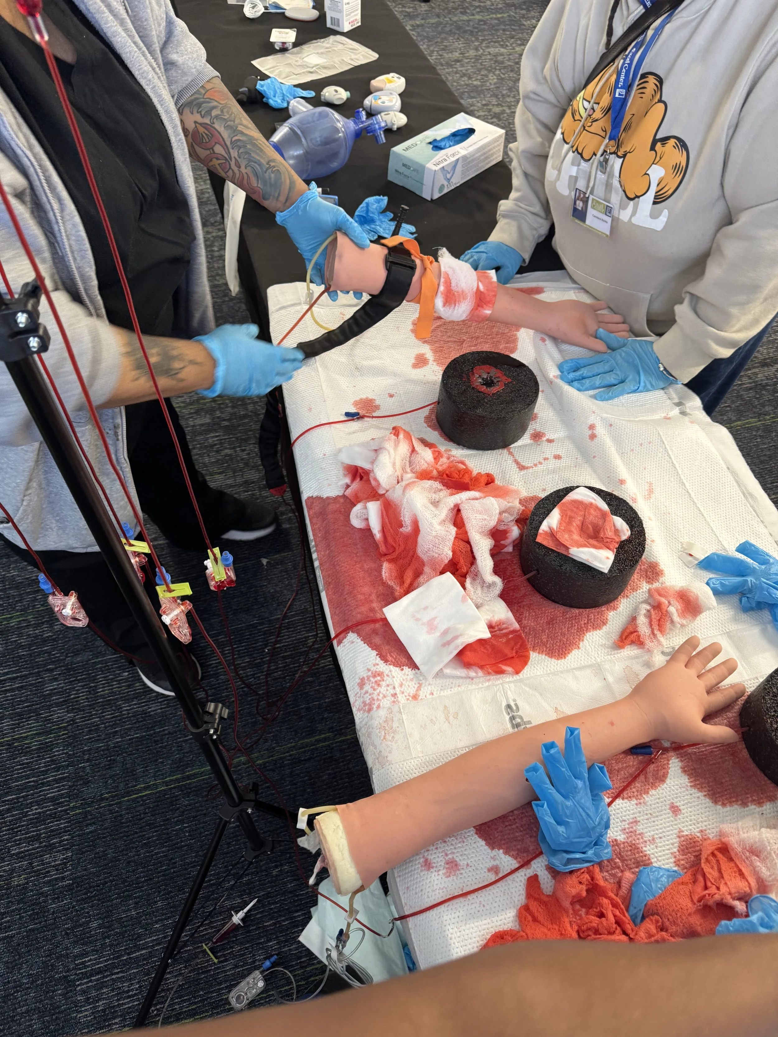 Medical personnel practicing on a simulated arm with blood and medical supplies nearby.