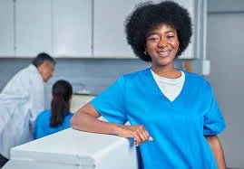 Young girl in blue scrubs smiling in a medical or clinical setting with healthcare workers in the background.