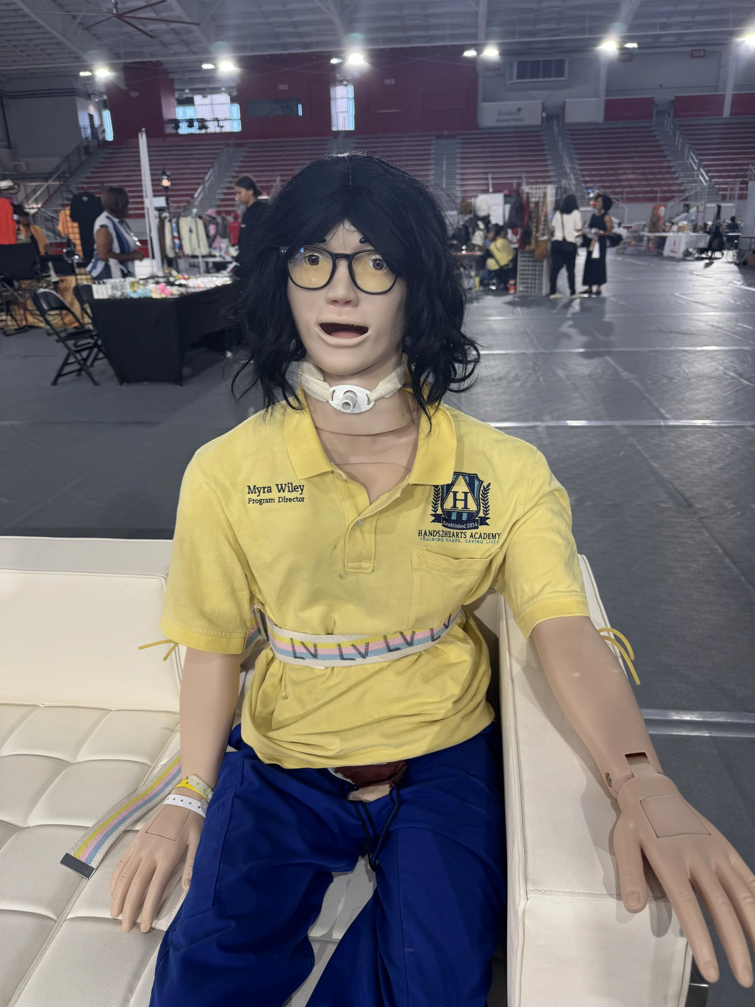 A mannequin dressed as a healthcare professional sitting on a white bench in a large indoor space with high ceilings, empty bleachers, and vendor booths in the background, wearing a yellow polo shirt with a logo and name tag, glasses, a neck brace, a