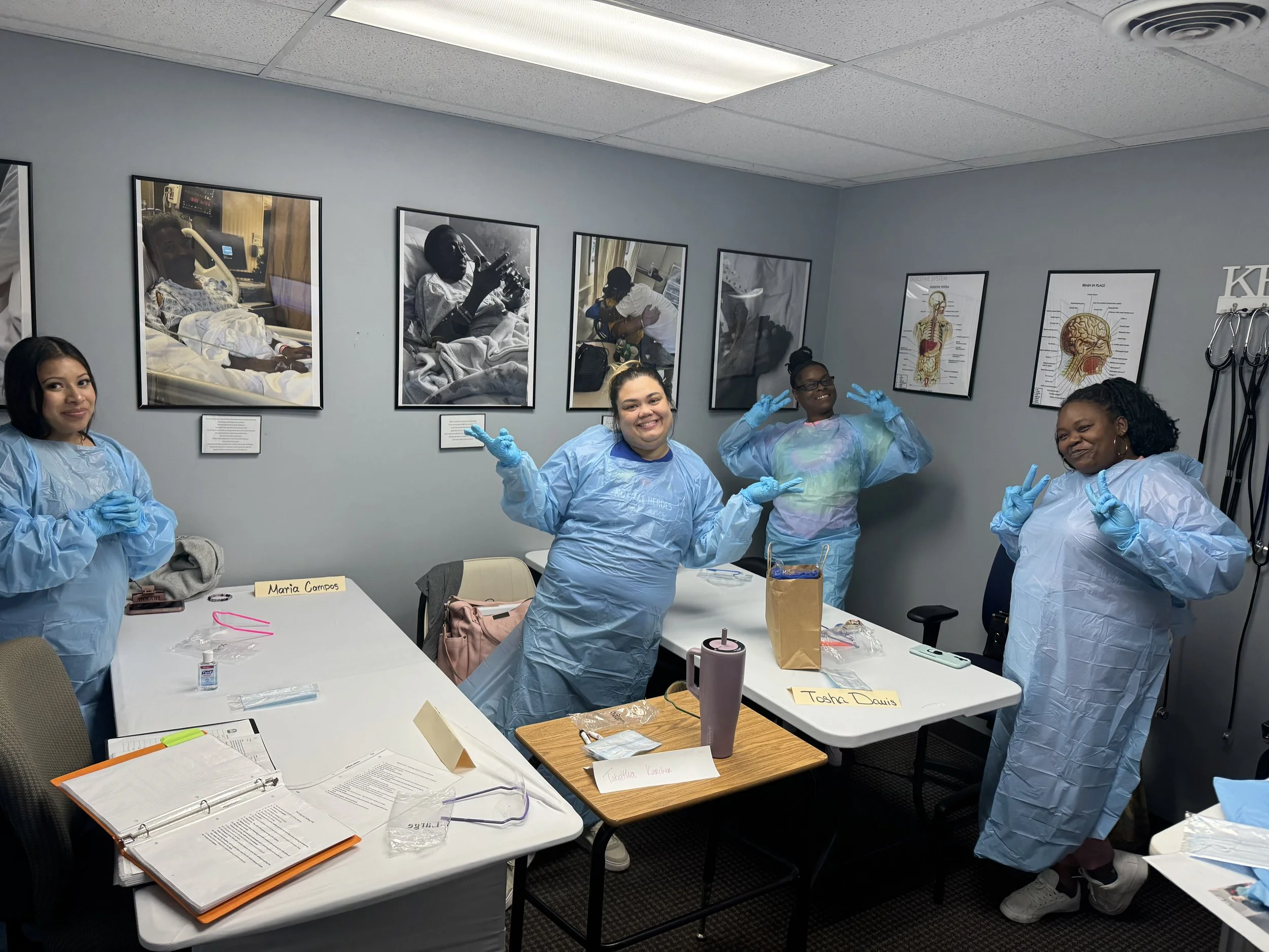 A group of women in medical scrubs and gloves posing in a conference room with medical posters and pictures of patients on the wall behind them. The women are smiling and making peace signs. The table in front of them has papers, notebooks, and perso