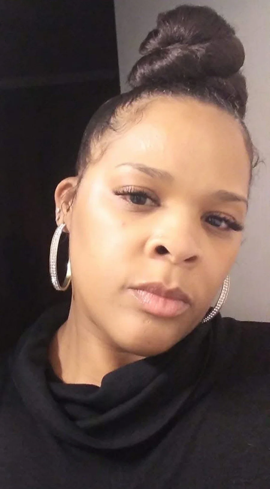 Close-up portrait of a young woman with a high bun hairstyle, hoop earrings, and a black top with a scarf or cowl neck. She has light makeup and a neutral expression.