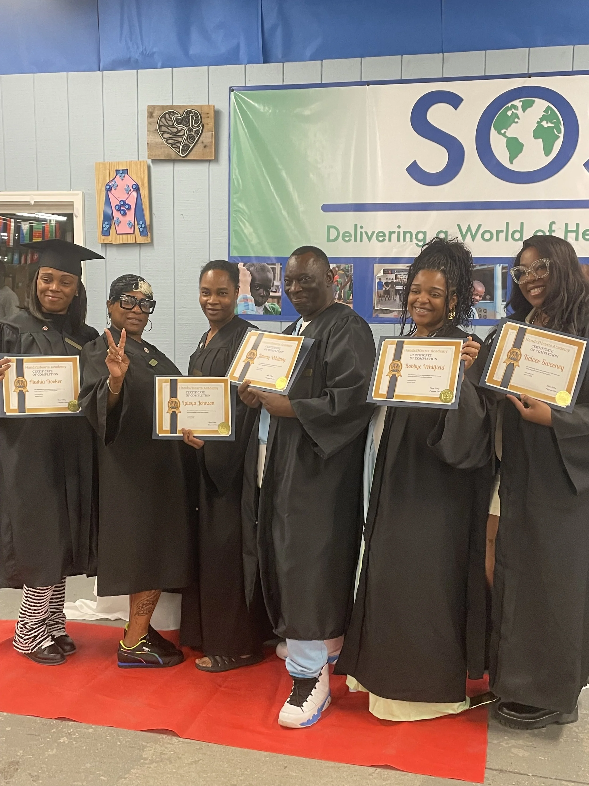 Group of people in graduation robes holding certificates, standing on a red carpet in front of a blue and white backdrop with logos and images, celebrating graduation.