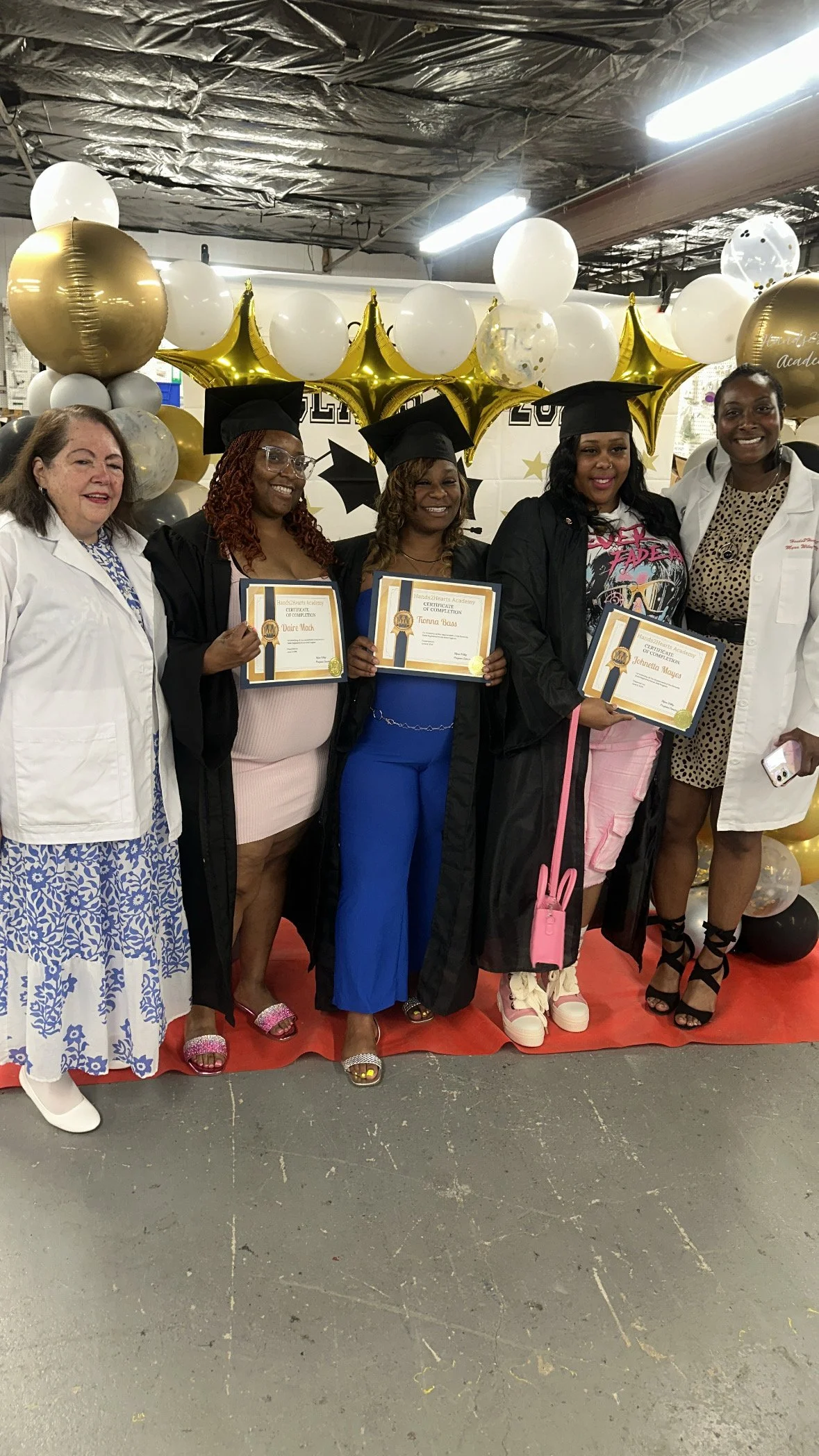 Celebration of graduation with diplomas, caps, and balloons, featuring formal and casual attire, in a decorated venue with balloons and a backdrop.