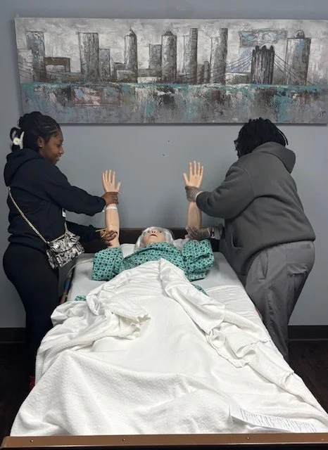 Two women are standing on either side of a hospital bed holding the hands of a person lying in the bed with a sheet covering their body. The person in bed is wearing a green hospital gown and has their hands raised in the air.