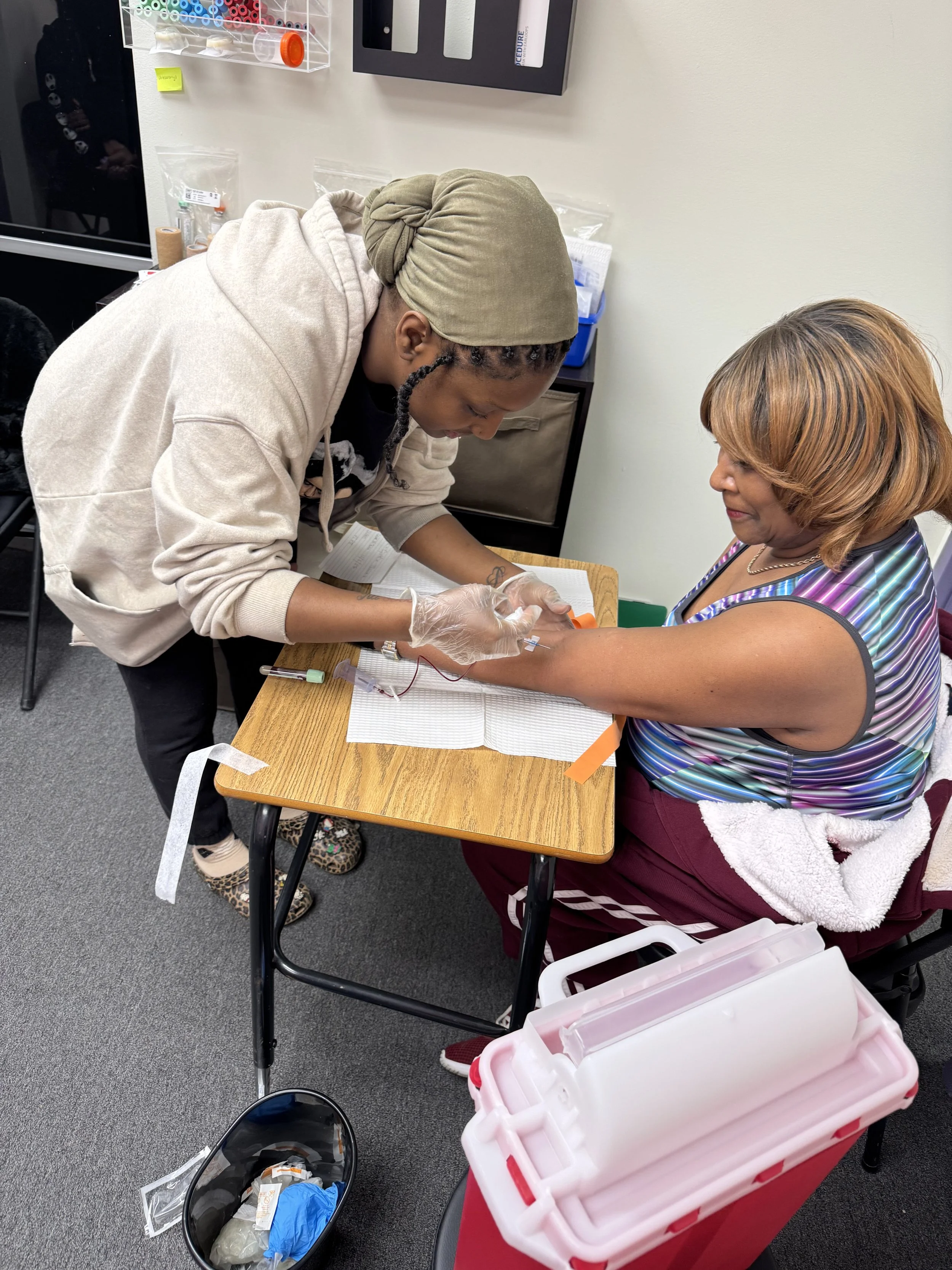 A healthcare professional and a woman receiving an injection in her arm during a medical procedure in a clinic room. The professional is dressed casually and wearing gloves, focusing on administering the injection, while the woman sits calmly. Medica