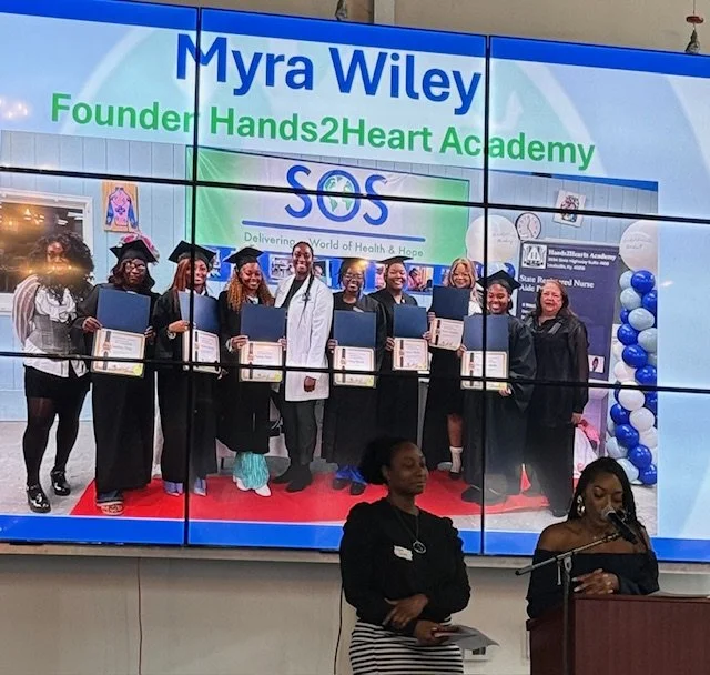 Group of graduates in caps and gowns holding diplomas at a graduation ceremony inside a building with a large screen displaying the name Myra Wiley, Hands2Heart Academy, and SOS, with decorations including balloons.