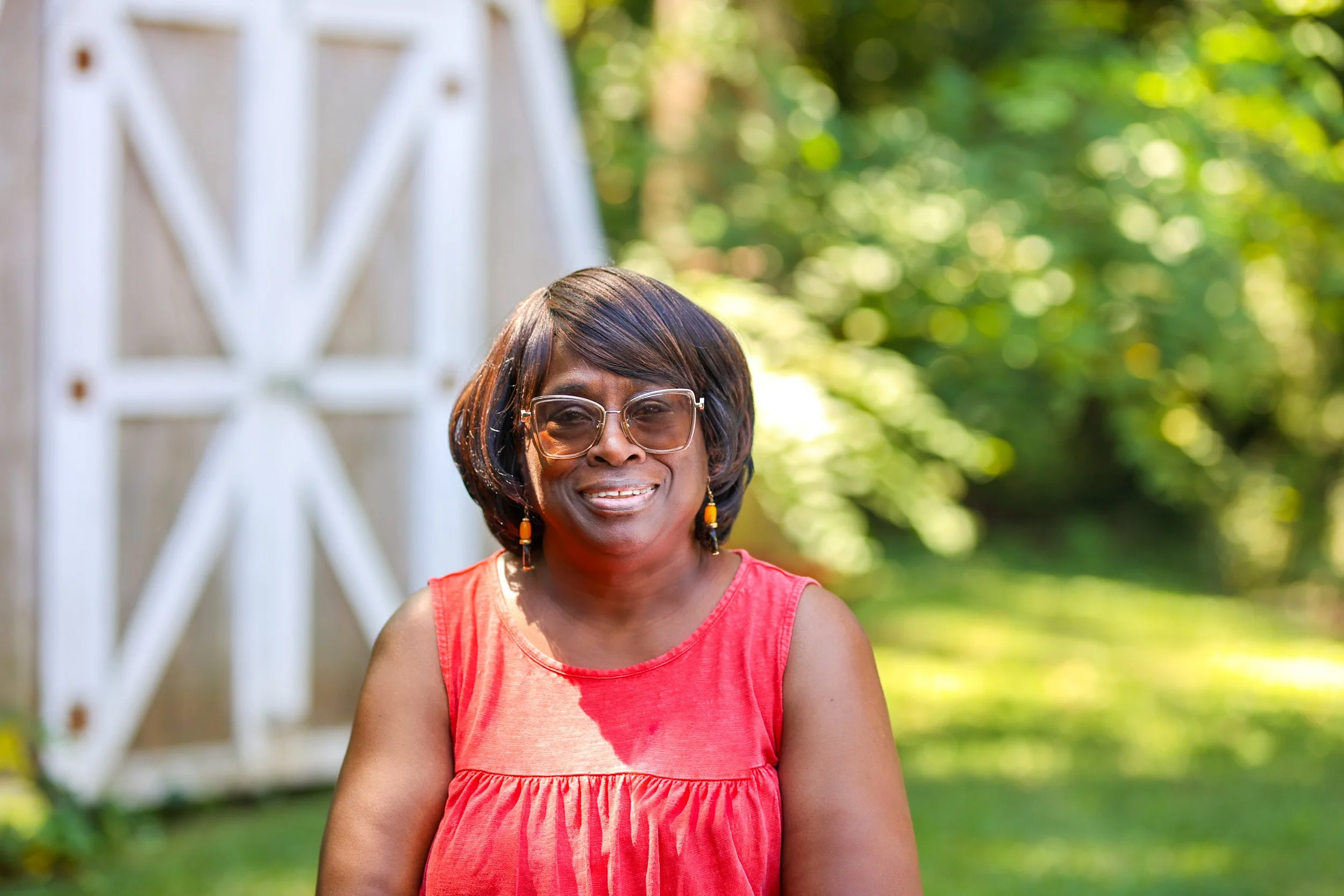 A middle-aged woman with short dark-hued hair, wearing glasses with a transparent frame, a sleeveless coral red top, and orange and black earrings, standing outside in front of a blurred background of green foliage and trees in sunlight.