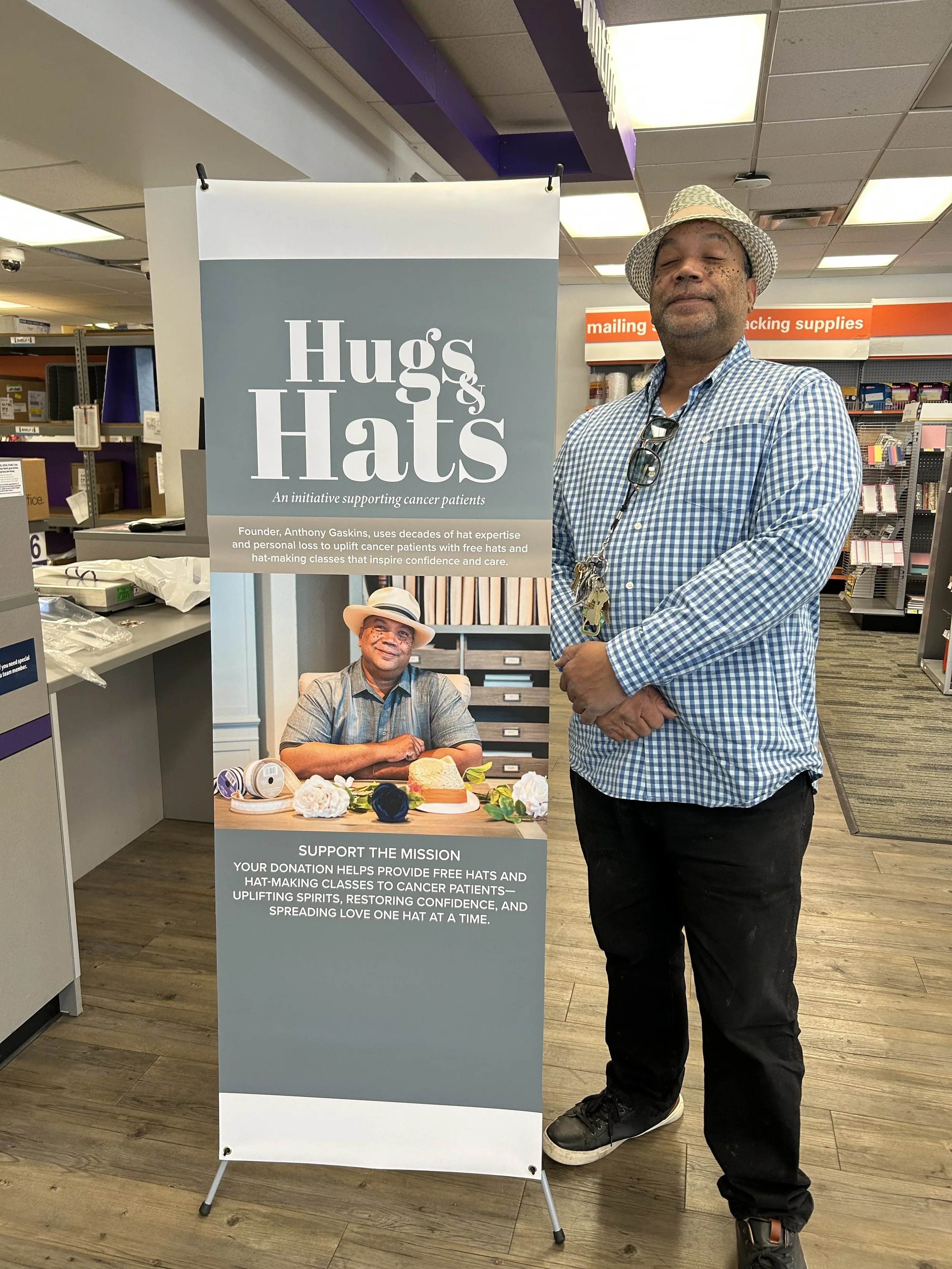 A man standing next to a banner in a store. The banner promotes "Hugs & Hats," an initiative for supporting cancer patients by providing free hats and hat-making classes to uplift their spirits and restore confidence.