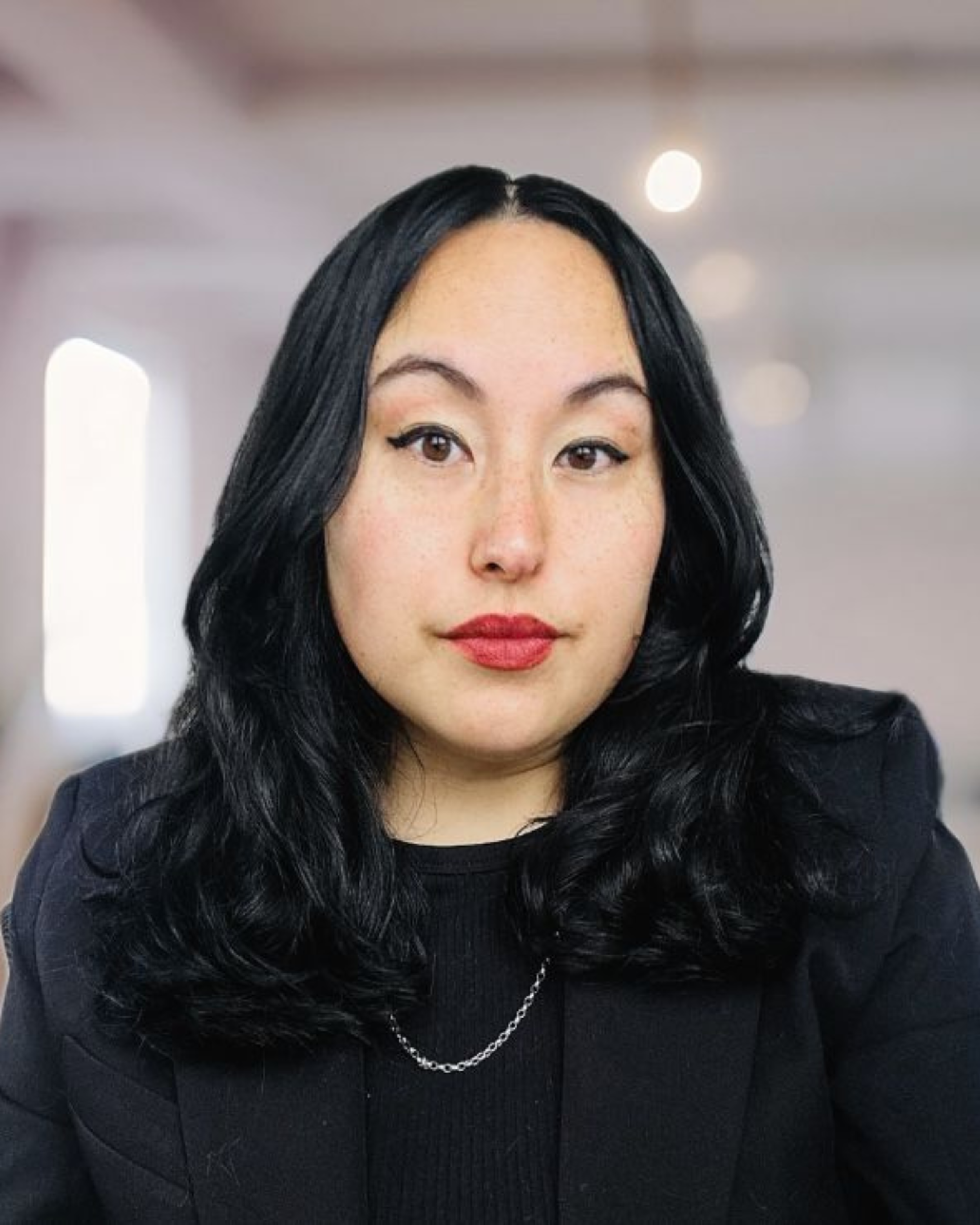 Close-up portrait of a woman with black hair, wearing a black blazer and a silver chain, in an indoor setting with blurred background.