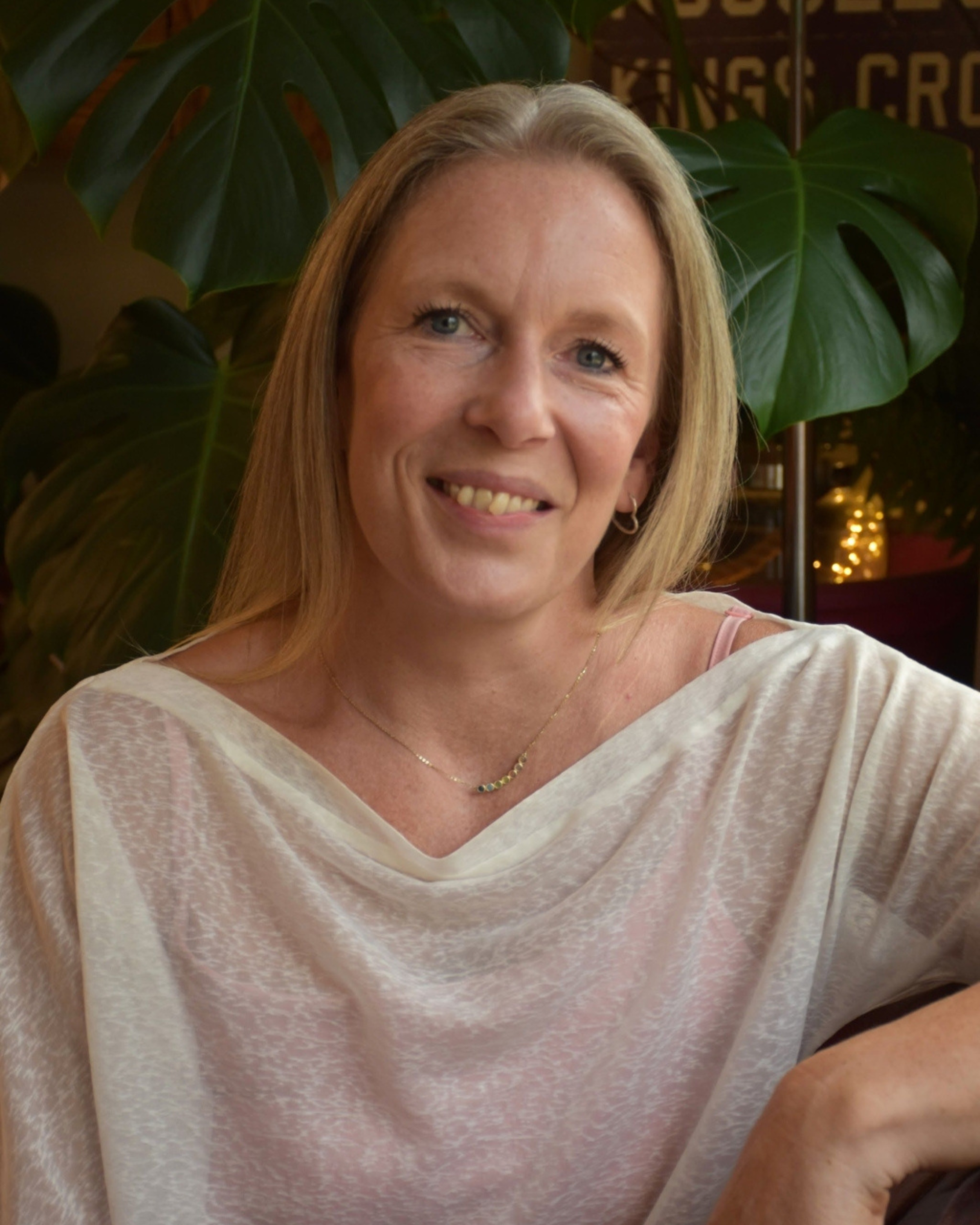 A woman with blonde hair smiling, sitting in front of large green leaves in an indoor setting with warm lighting.
