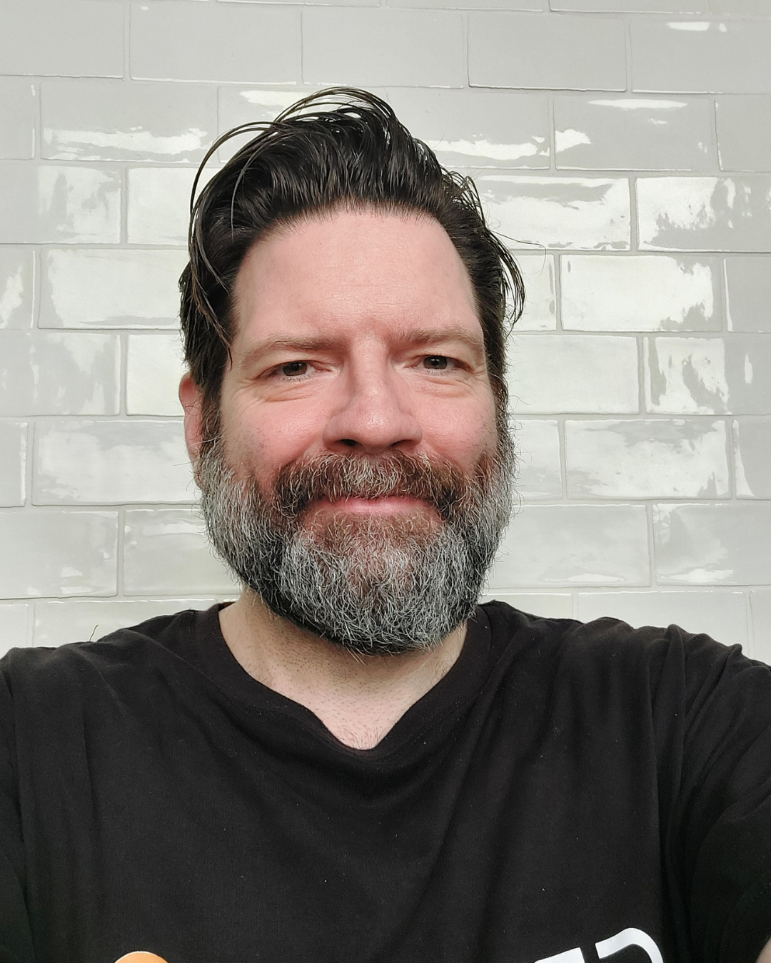 A man with dark, slicked-back hair and a beard, wearing a black shirt, looking directly into the camera against a plain white background.