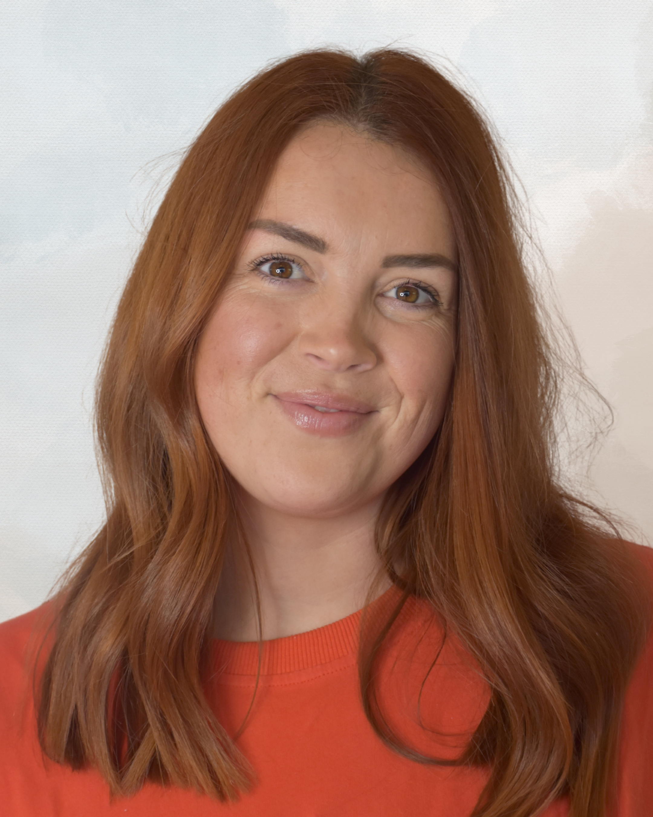 A woman with long, red hair wearing an orange top, smiling softly, against a neutral background.