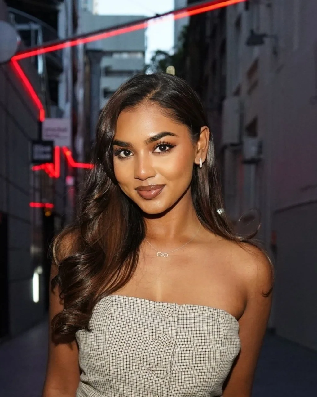 A young woman with long styled hair, wearing a strapless checkered top, jewelry including a necklace and earrings, standing on a city street during twilight with red neon lights in the background.