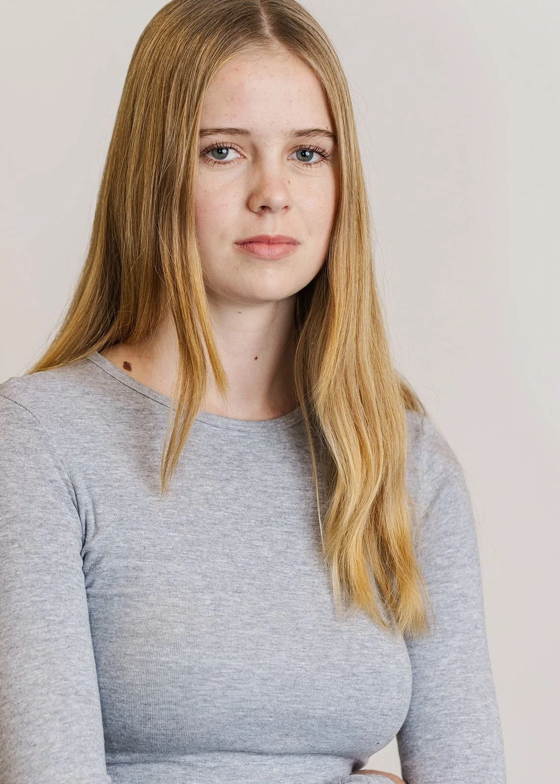 A young woman with long red hair and blue eyes looking at the camera, wearing a light gray shirt, against a plain background.