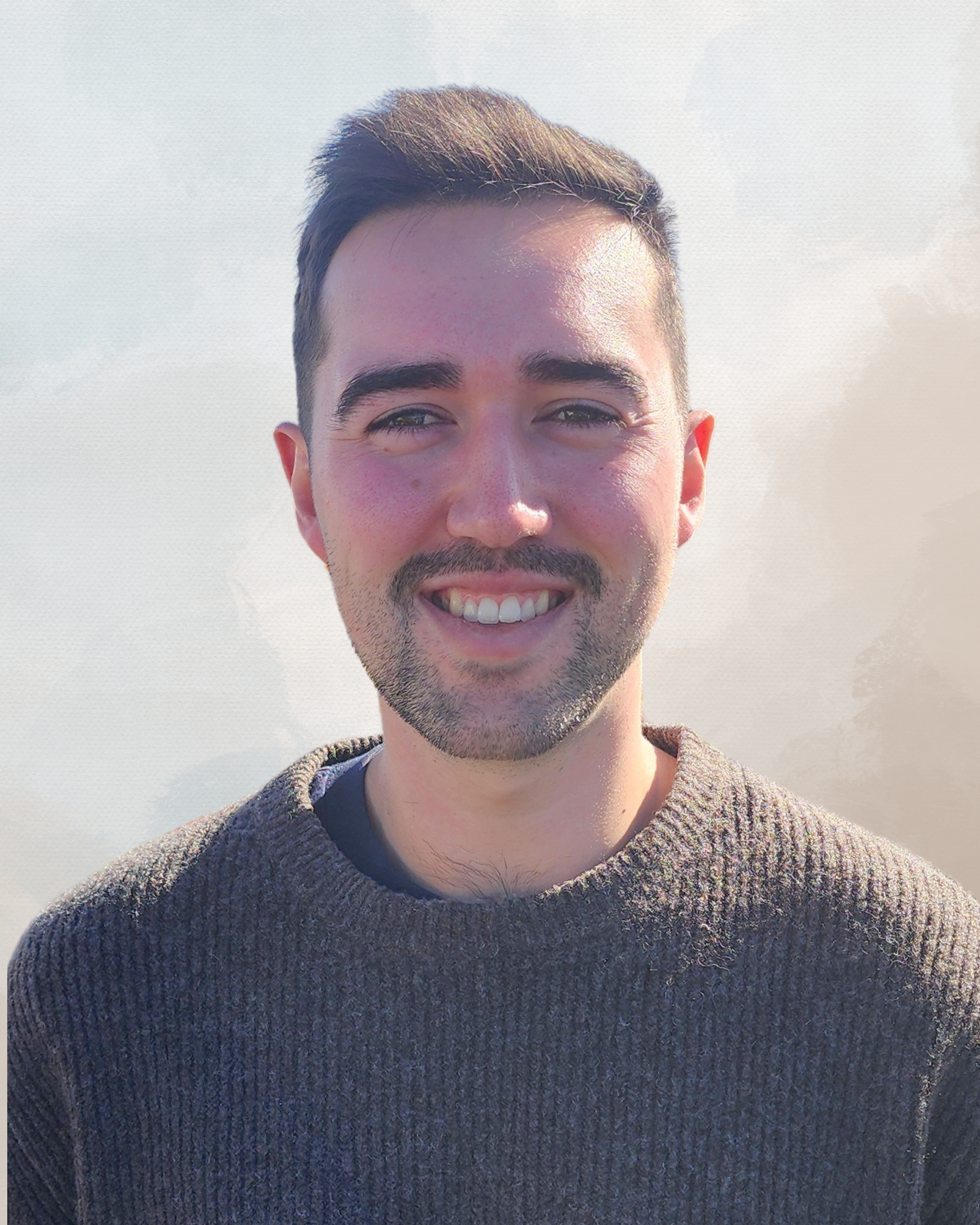 A smiling man outdoors with sunlight on his face, wearing a brown sweater against a cloudy sky background.