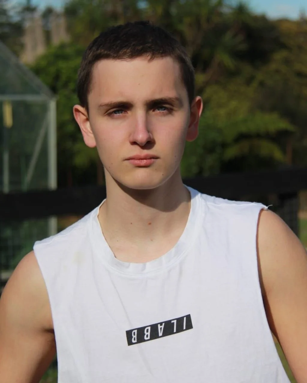 A young man with short dark hair and blue eyes standing outdoors in front of green trees, wearing a white sleeveless shirt with a black and white label that says 'ILABB'.
