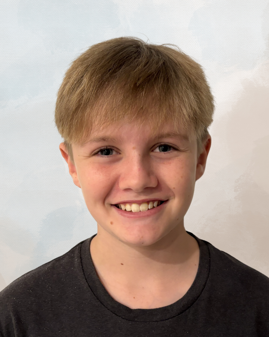 A smiling young boy with short light brown hair, blue eyes, and freckles, wearing a black t-shirt against a neutral background.