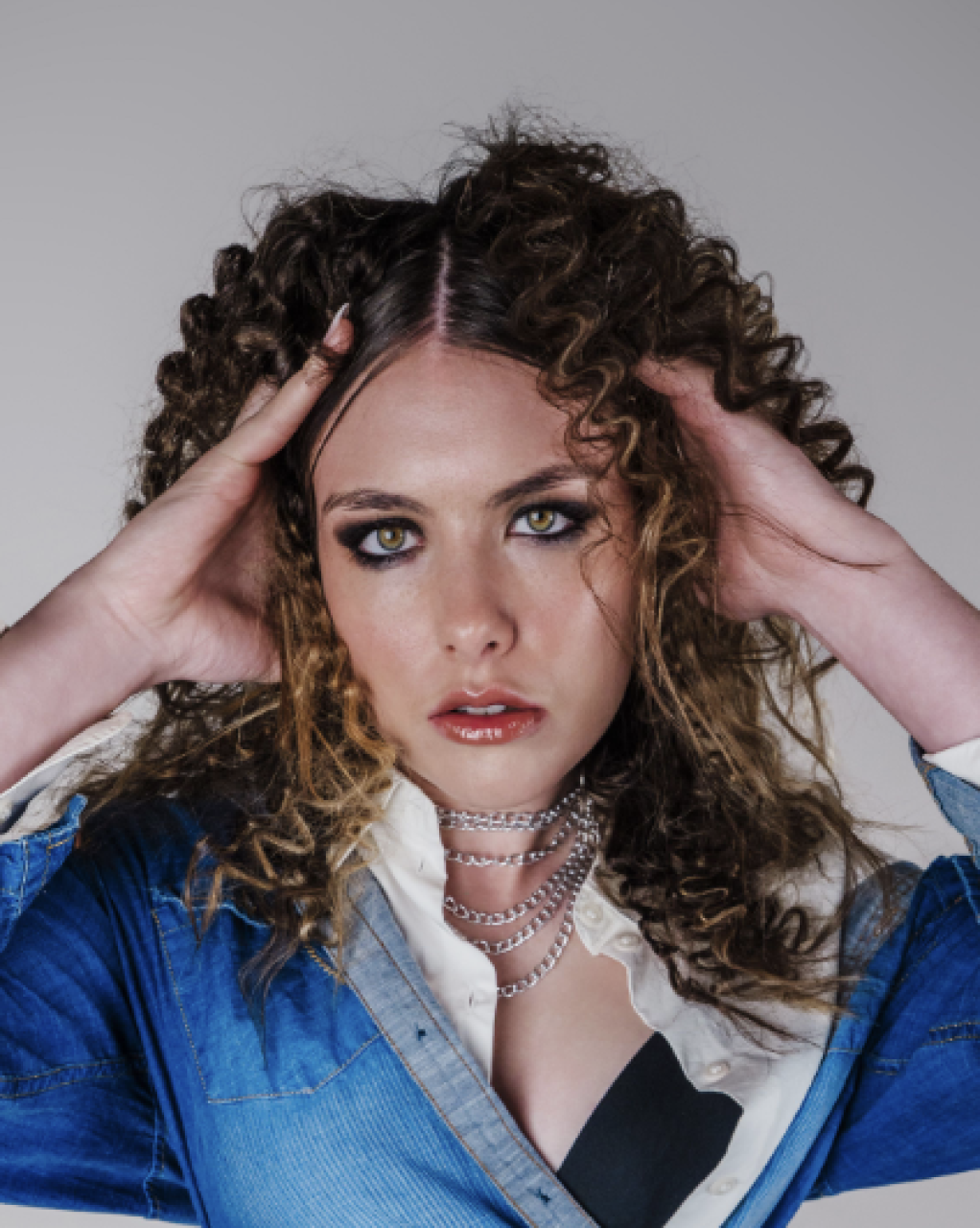 A woman with curly brown hair and striking makeup, wearing a denim jacket, layered silver necklaces, and a white blouse, holding her head with both hands.
