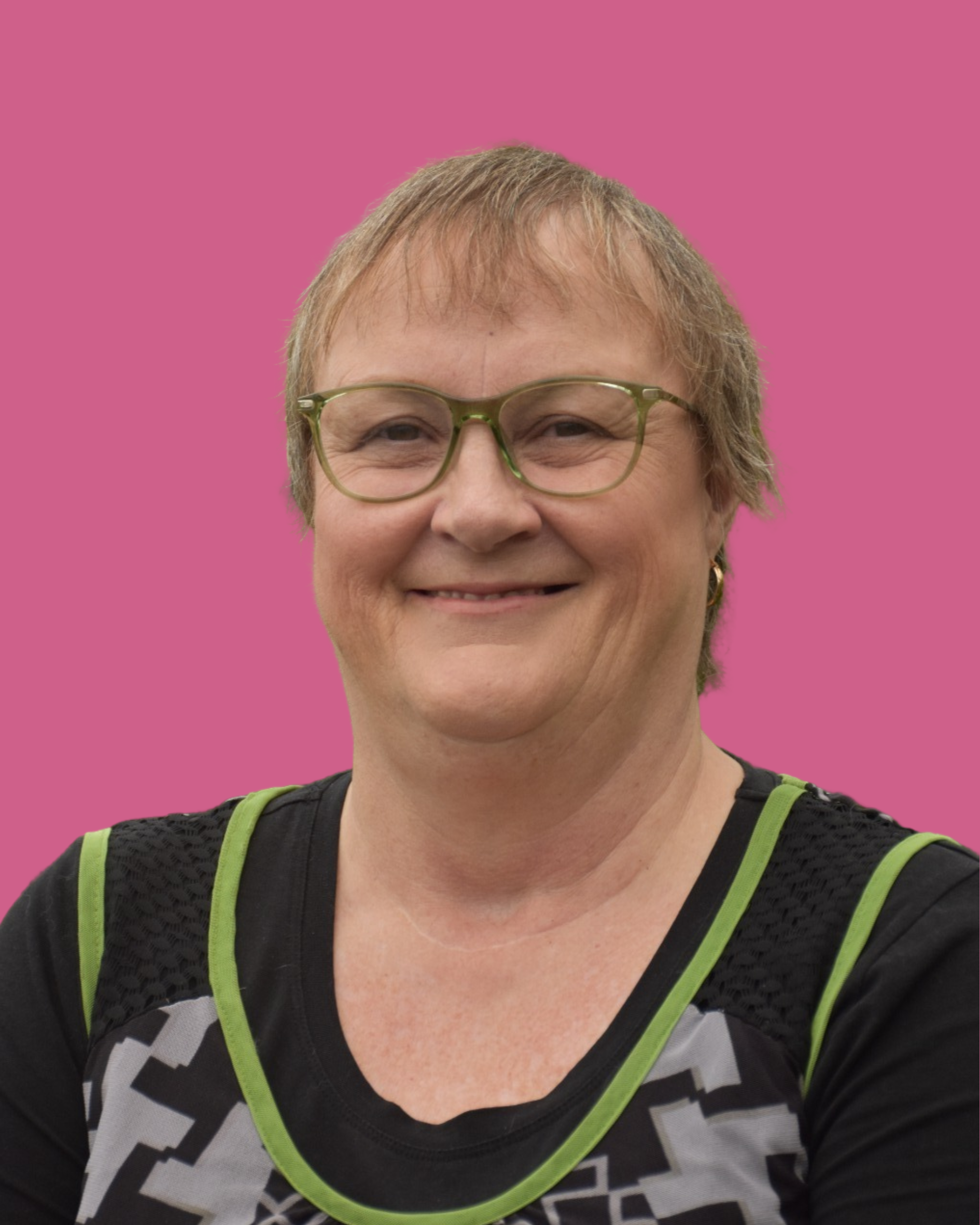 A woman with short light brown hair, wearing glasses, a black athletic shirt with green accents, and earrings, smiling against a pink background.