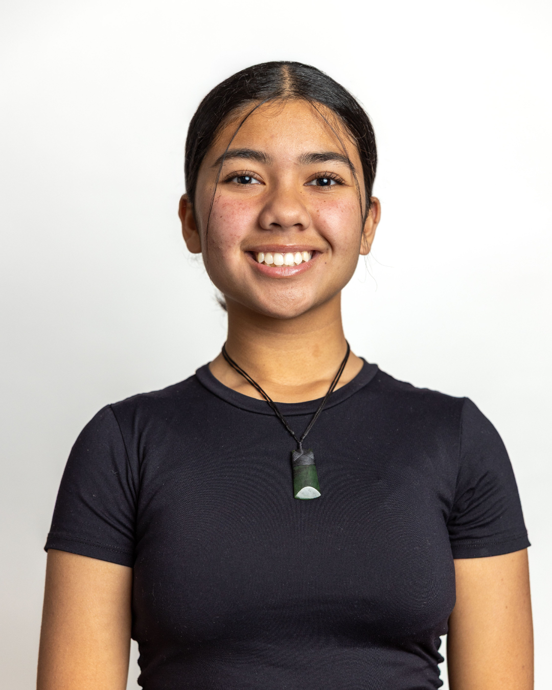 A young woman with dark hair in a ponytail, smiling, wearing a black t-shirt and a black necklace with a green and white pendant, standing against a plain white background.