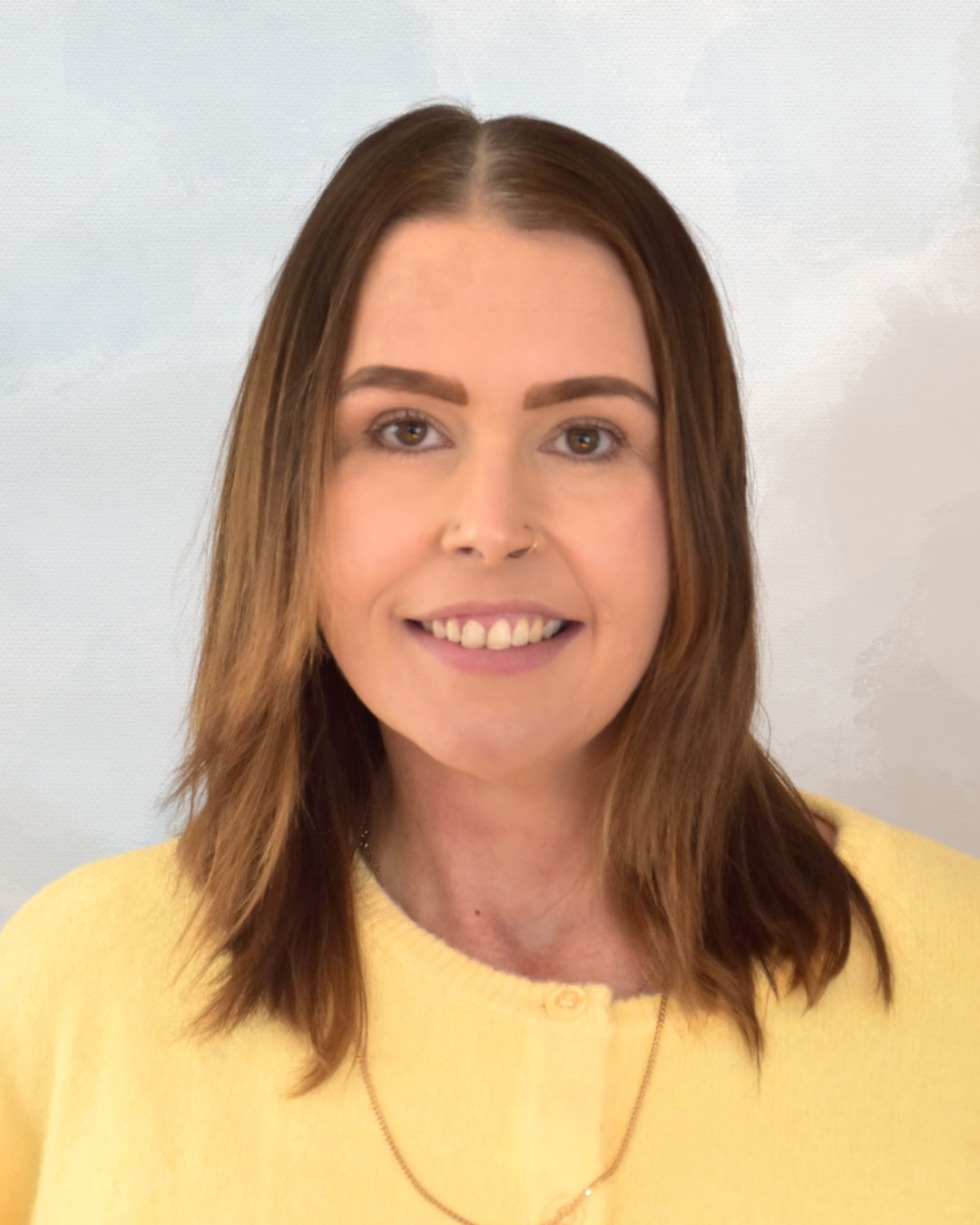A woman with shoulder-length brown hair, light skin, and brown eyes, smiling, wearing a yellow top and a gold necklace, standing against a light background.