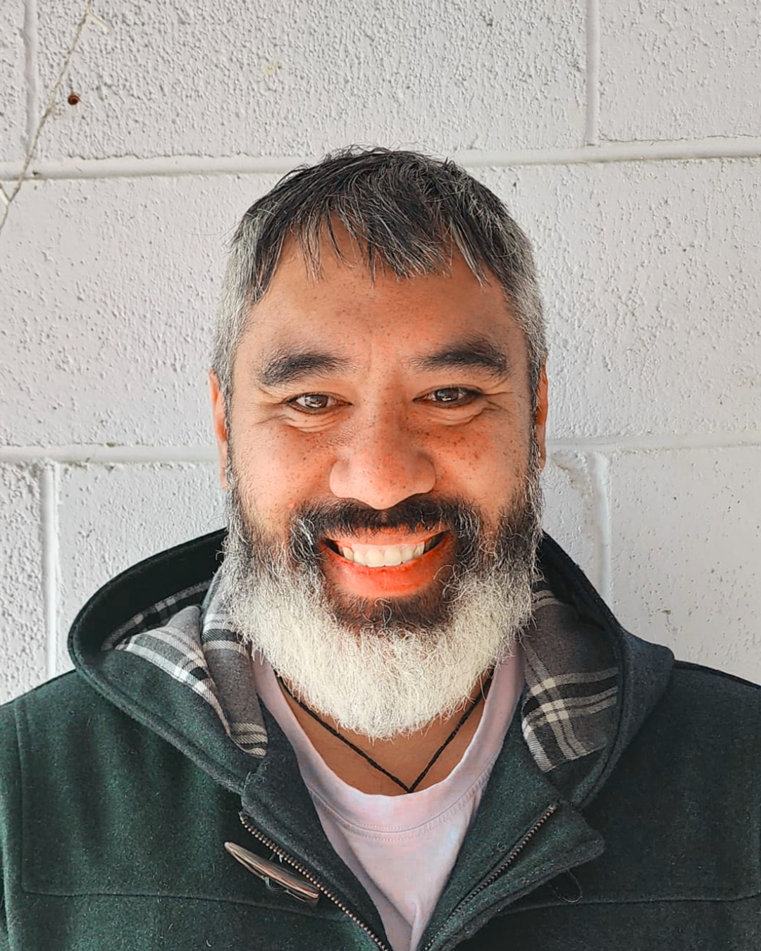 A smiling man with gray hair and beard wearing a dark zip-up jacket against a white brick wall.