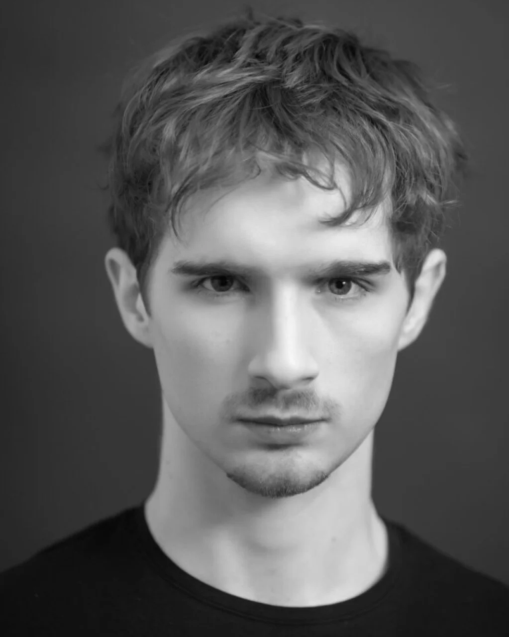 Black and white portrait of a young man with styled, wavy hair, intense gaze, light skin, and slight facial hair, wearing a black top against a dark background.