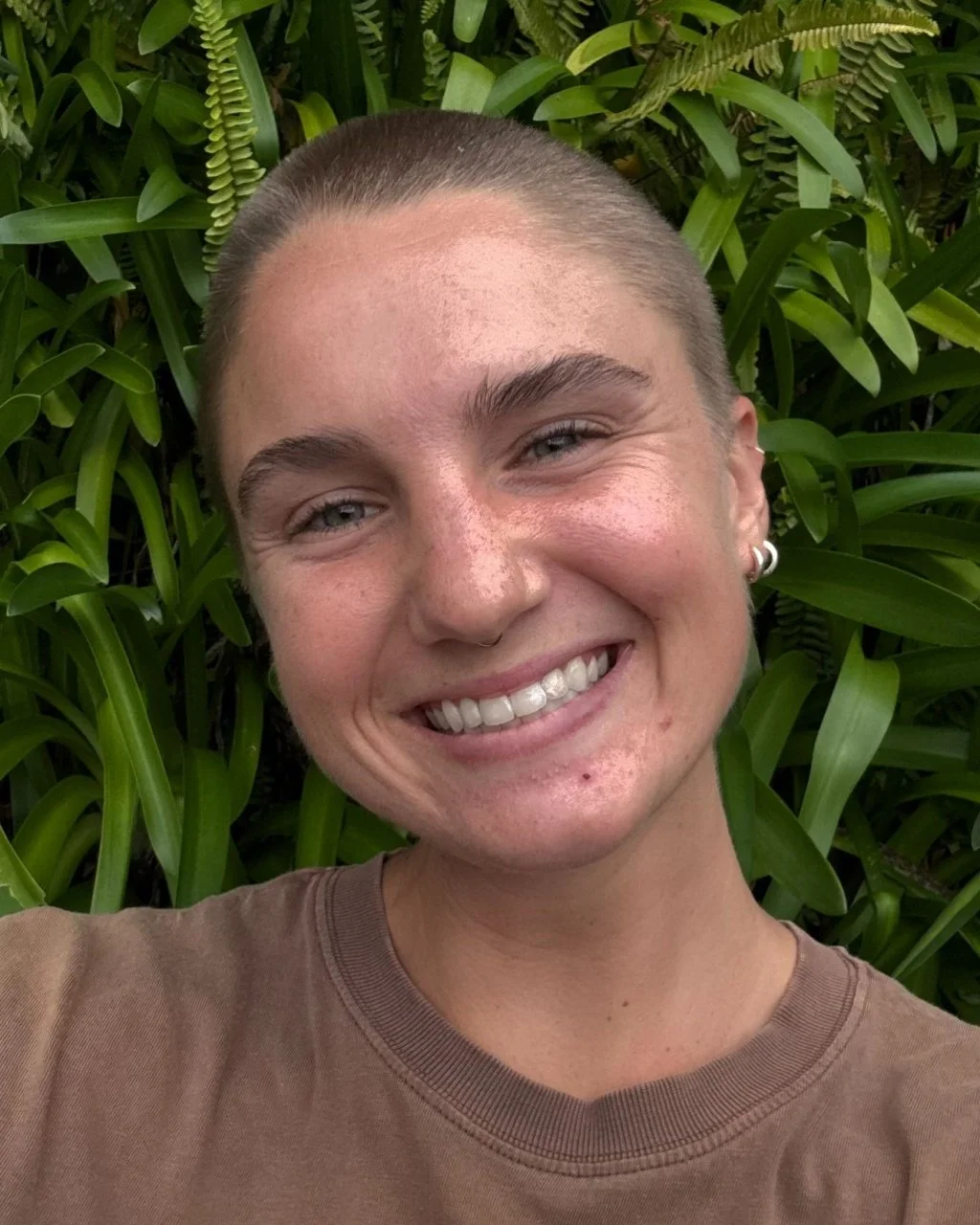 Close-up of a smiling person with short hair and earrings, standing in front of lush green plants.