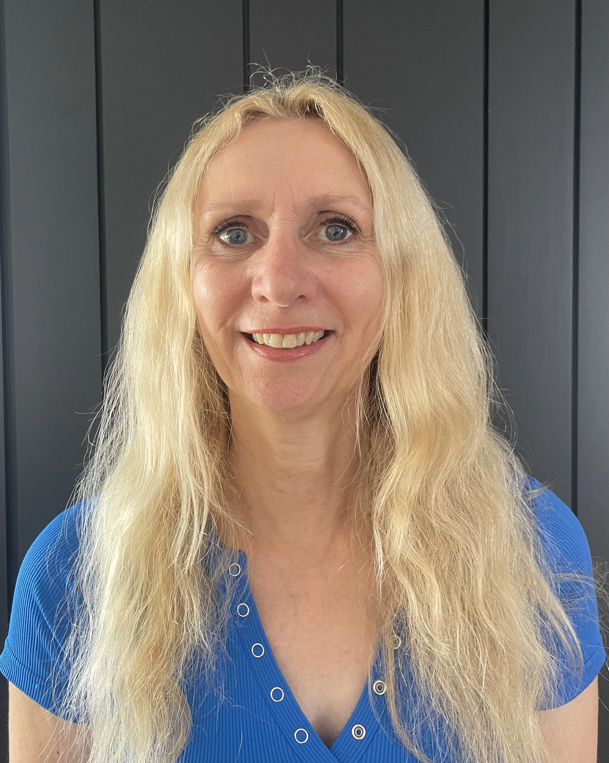 A woman with long blonde hair, blue eyes, and a bright smile wearing a blue top stands in front of a dark paneled wall.