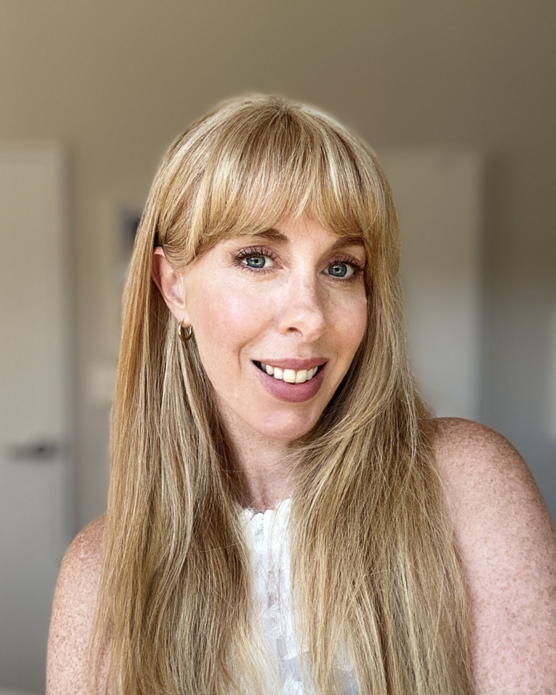 A woman with long, wavy blonde hair and blue eyes smiling indoors.