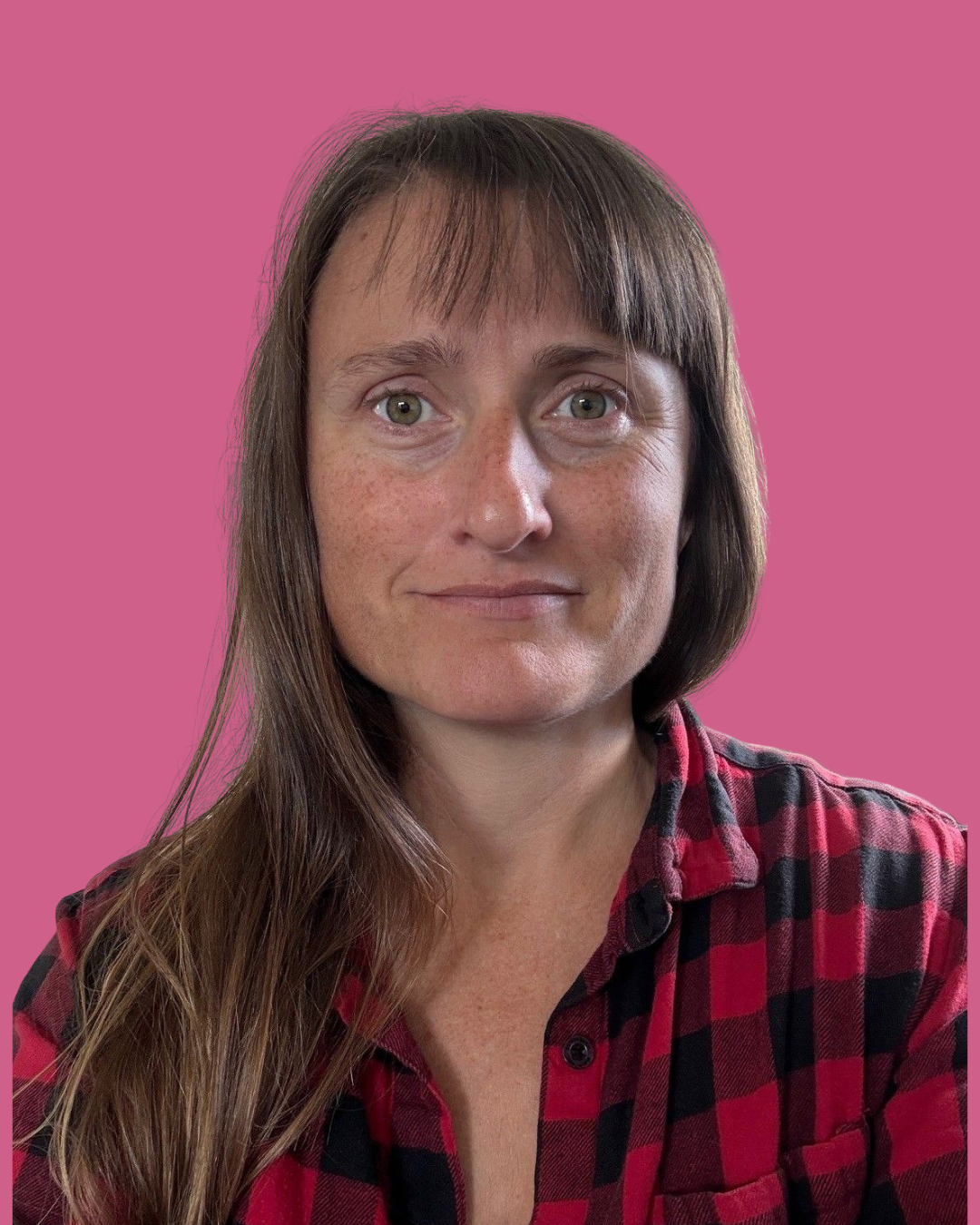 A woman with long brown hair, green eyes, and freckles, wearing a red and black plaid shirt, standing against a pink background.