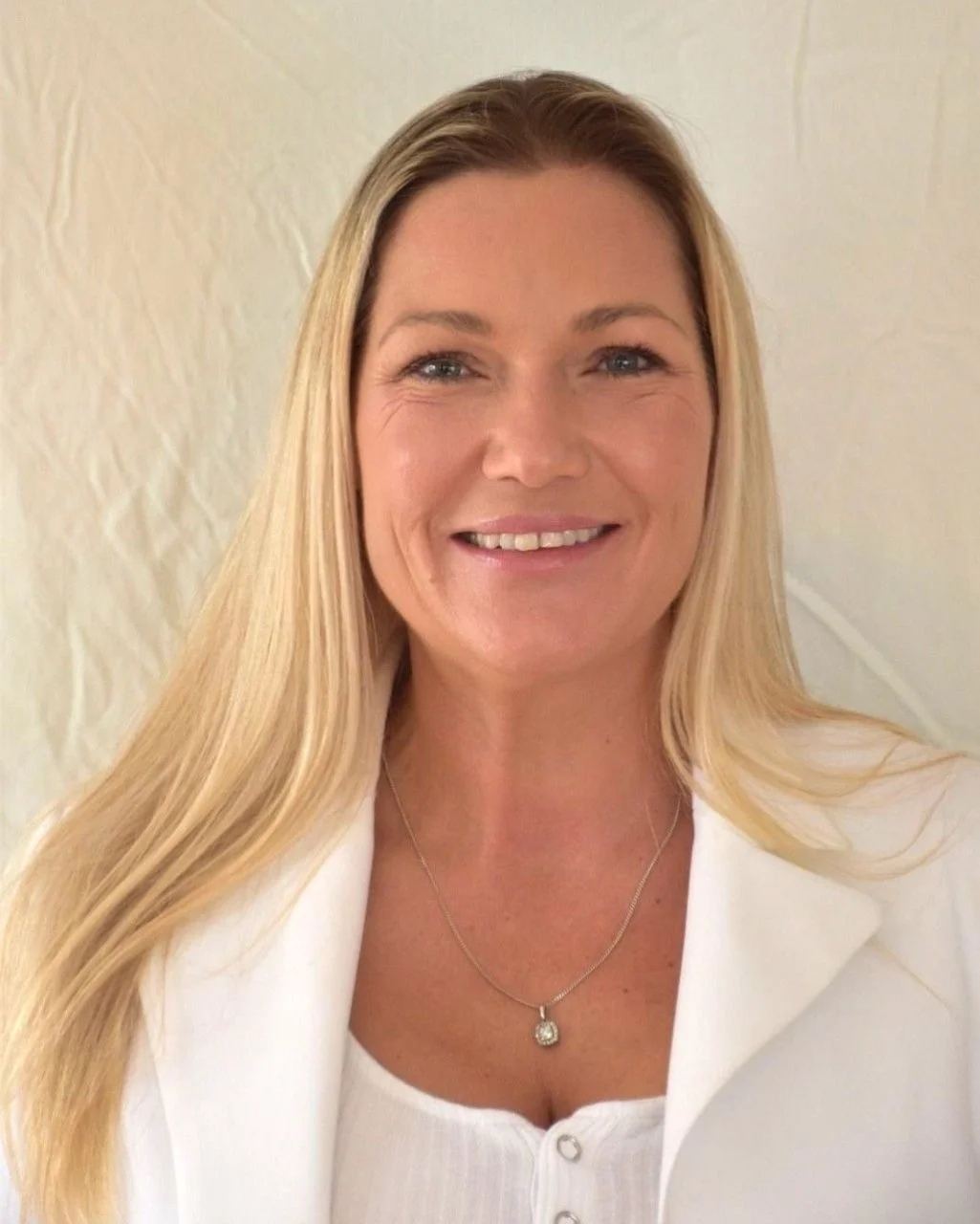 A smiling woman with long blonde hair, wearing a white blazer and a necklace, standing against a light background.