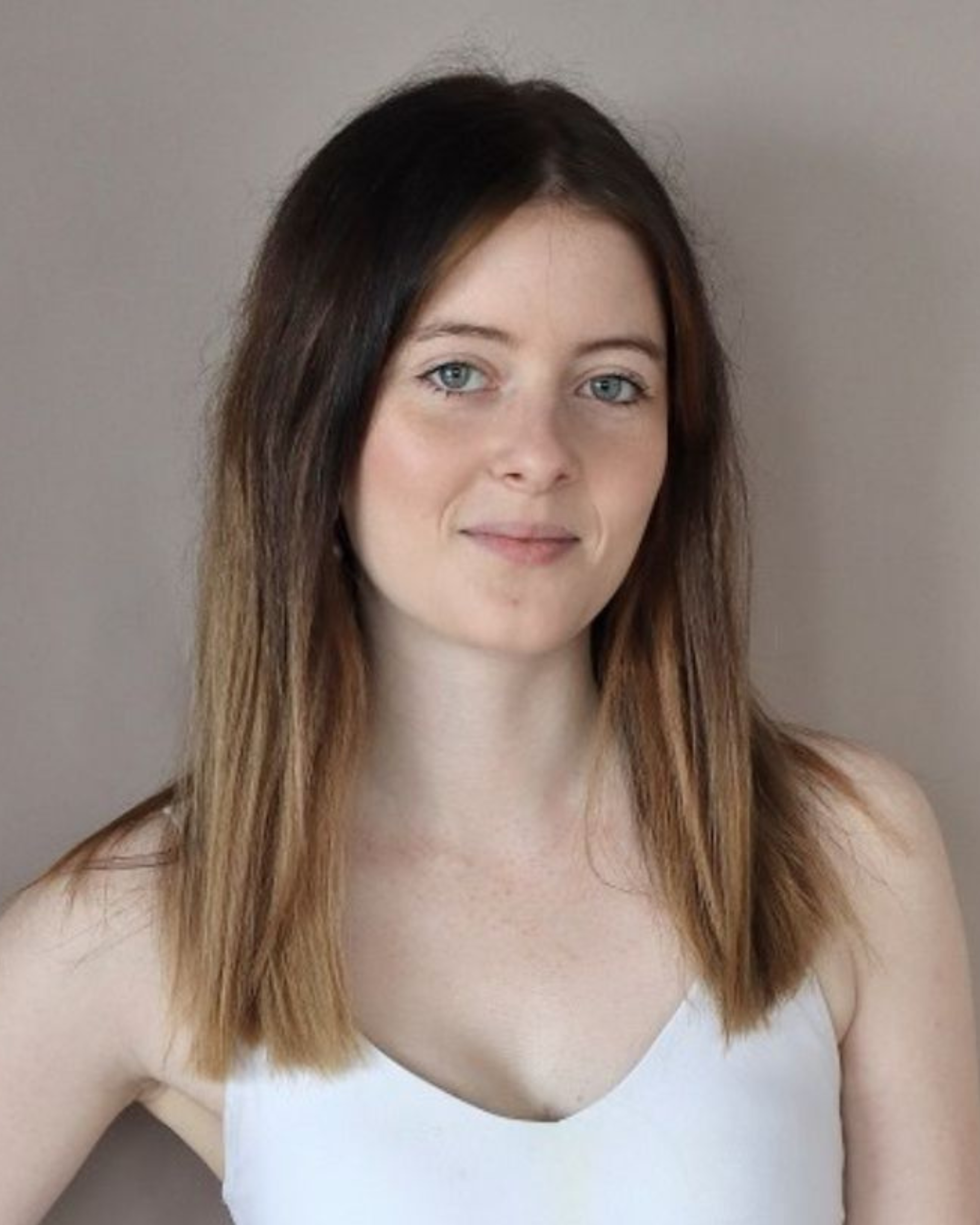A woman with shoulder-length brown hair, blue eyes, and fair skin wearing a white sleeveless top, standing against a plain gray background.