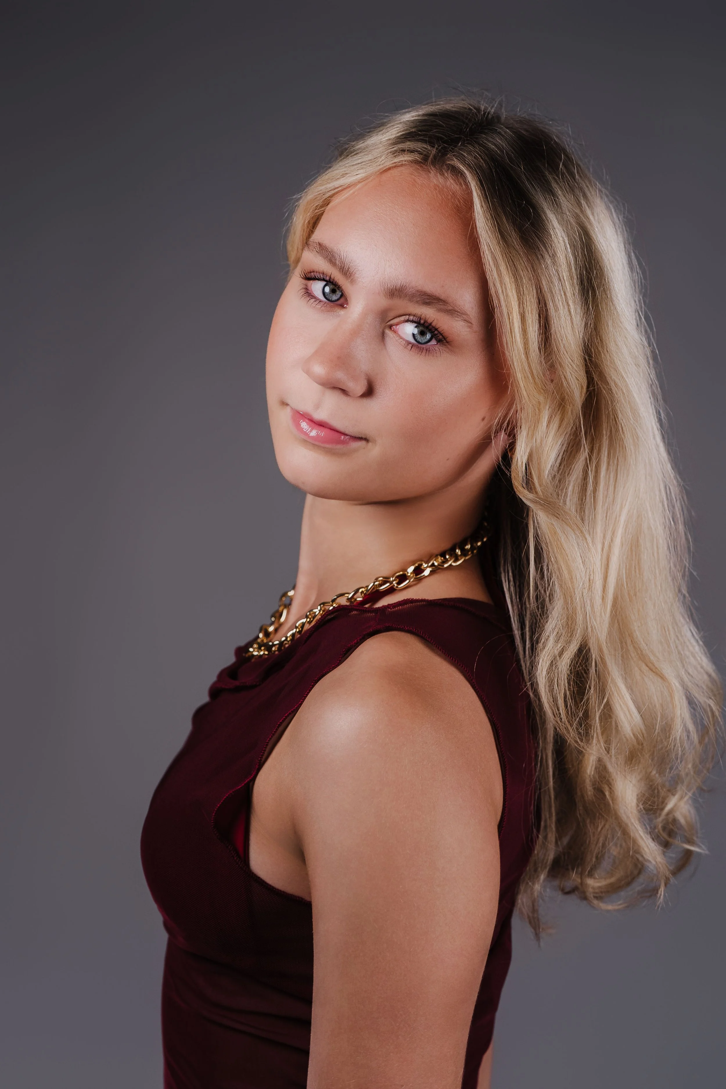 Portrait of a young woman with blonde wavy hair, blue eyes, wearing a maroon sleeveless top and a gold chain necklace, against a gray background.
