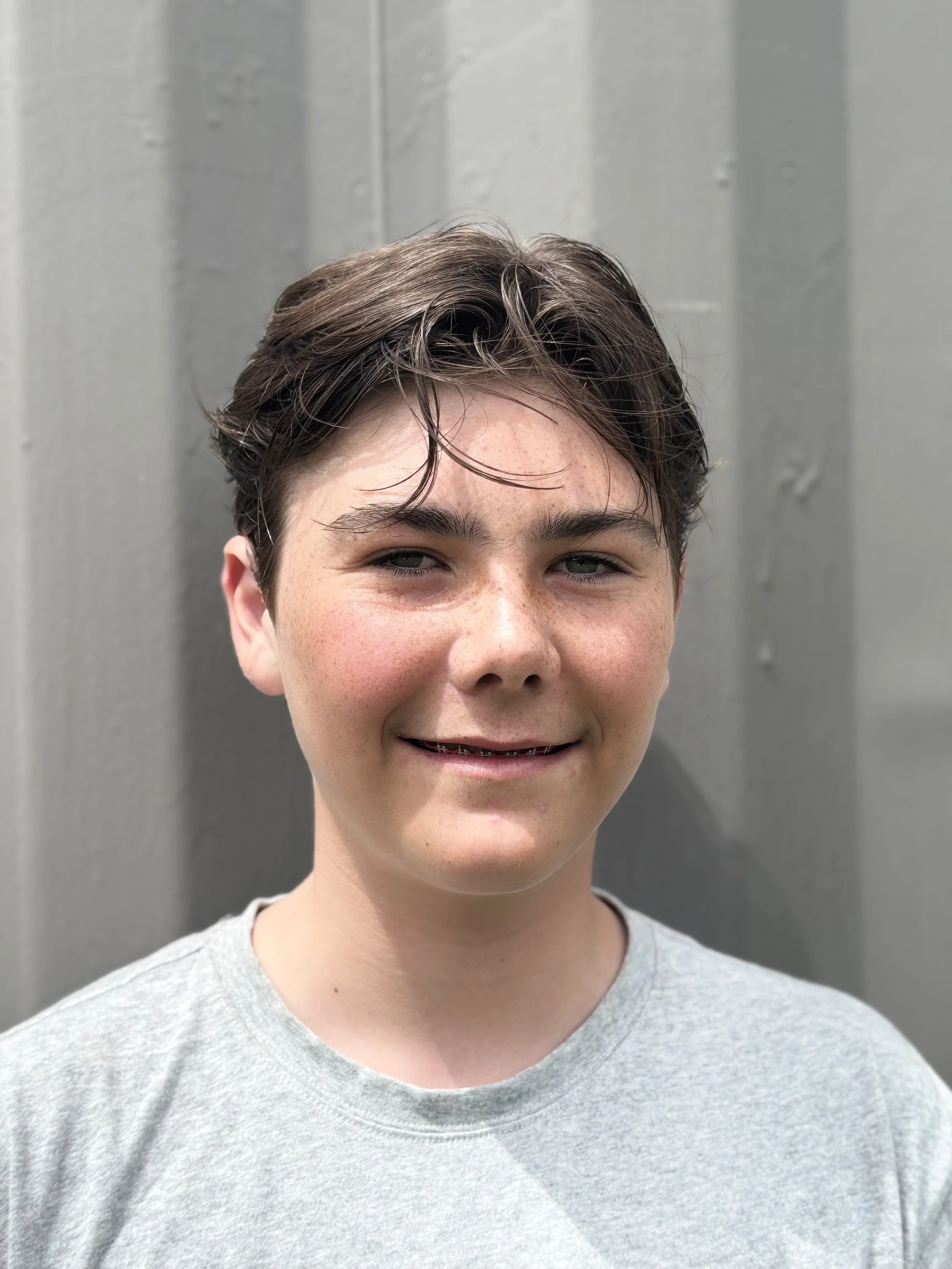 Close-up image of a smiling young person with short, dark hair in front of a gray wooden wall.