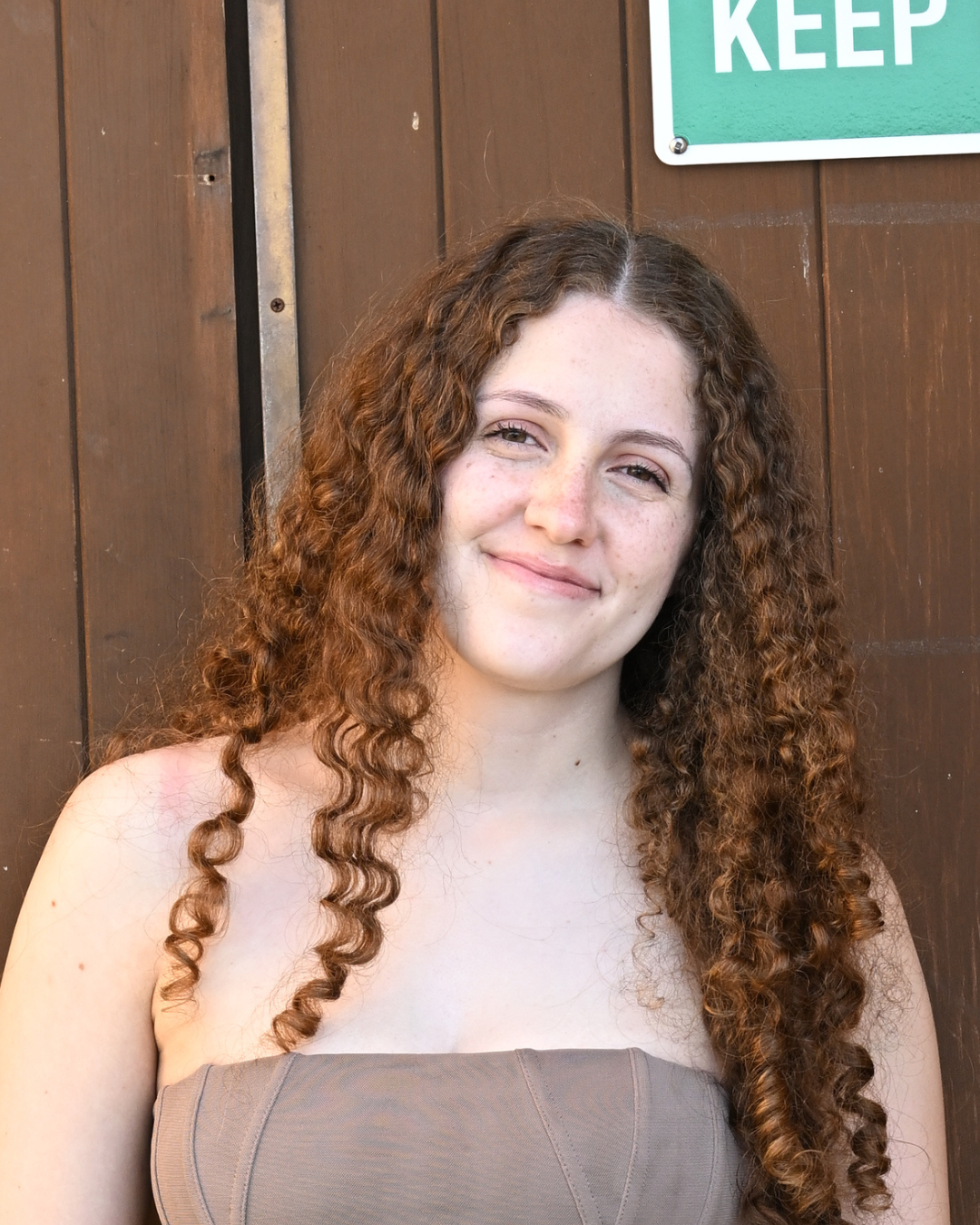 A young woman with long, curly red hair smiling in front of a wooden wall with a green sign.