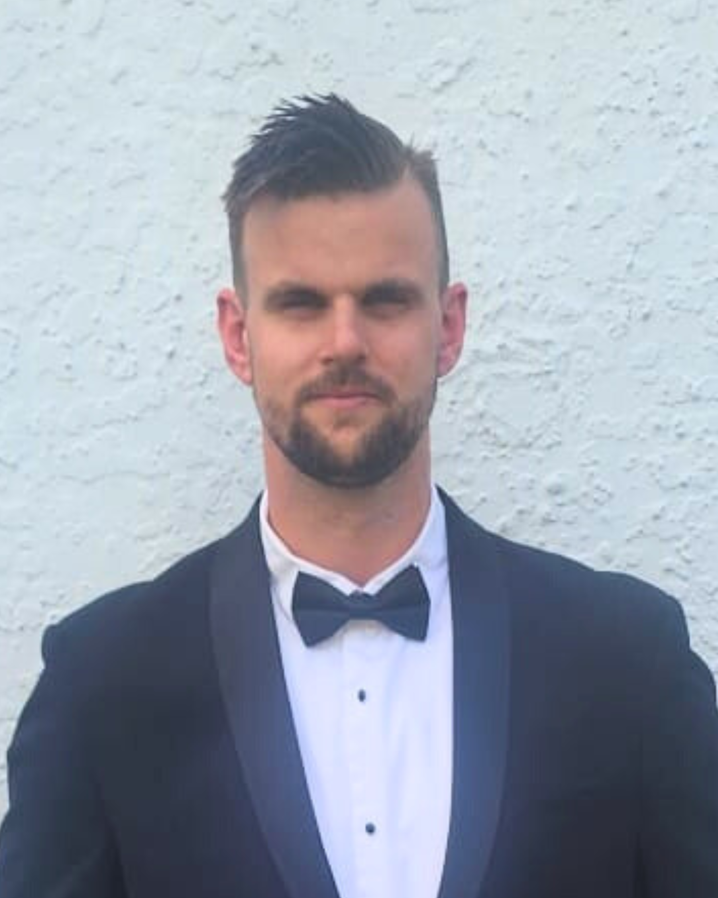 A young man with styled dark hair, a beard, and a serious expression, dressed in a tuxedo with a bow tie, standing in front of a white textured wall.