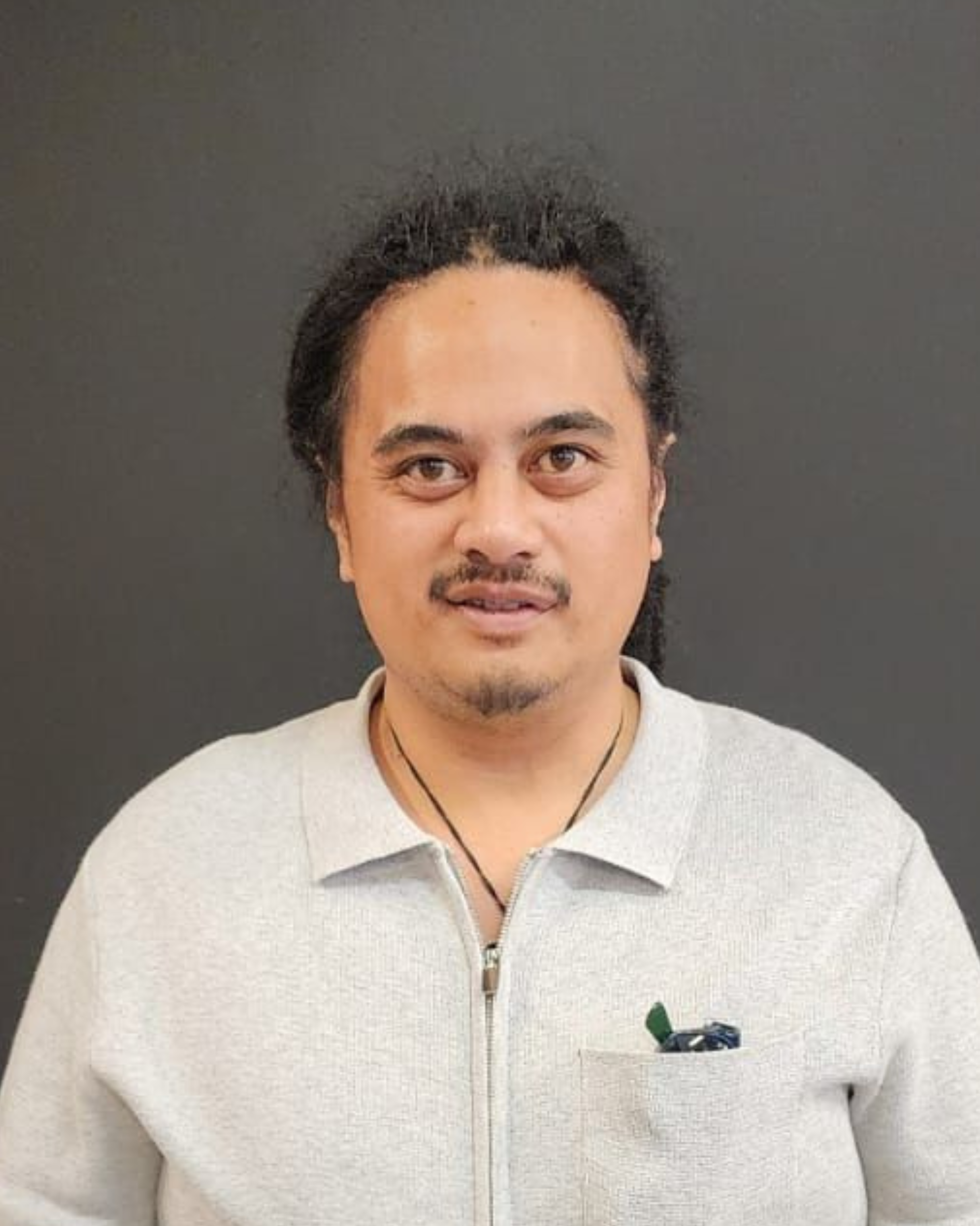 A man with curly black hair, mustache, and goatee, wearing a light gray zip-up sweater, standing against a dark background.