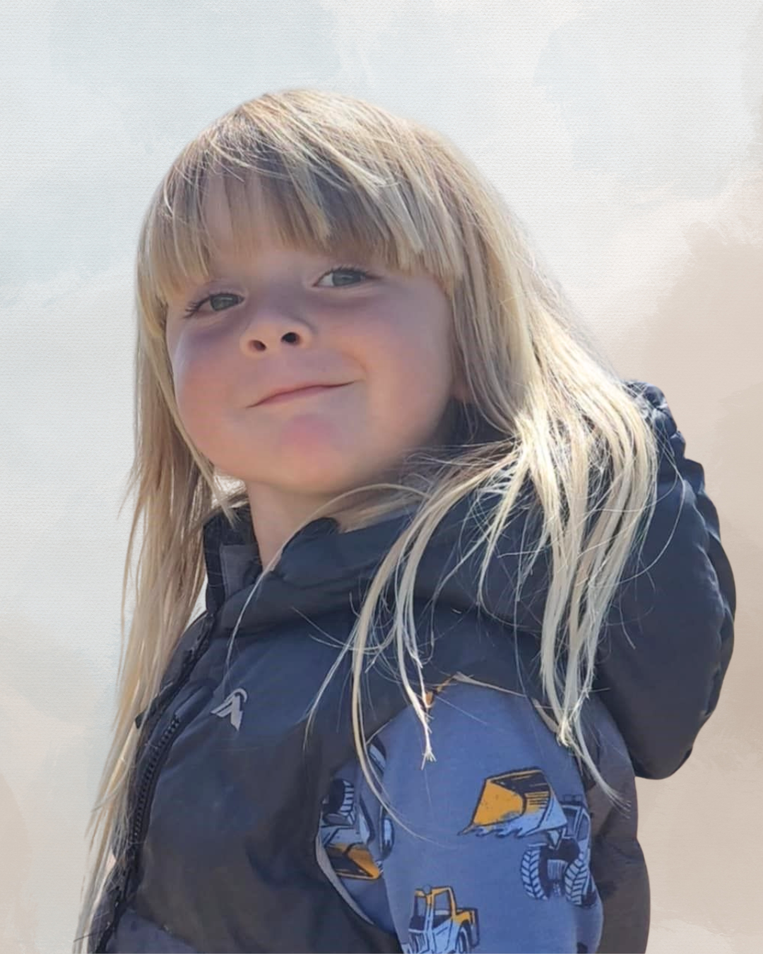 Young blonde girl outdoors, smiling, wearing a black jacket with a yellow and blue construction vehicle pattern.