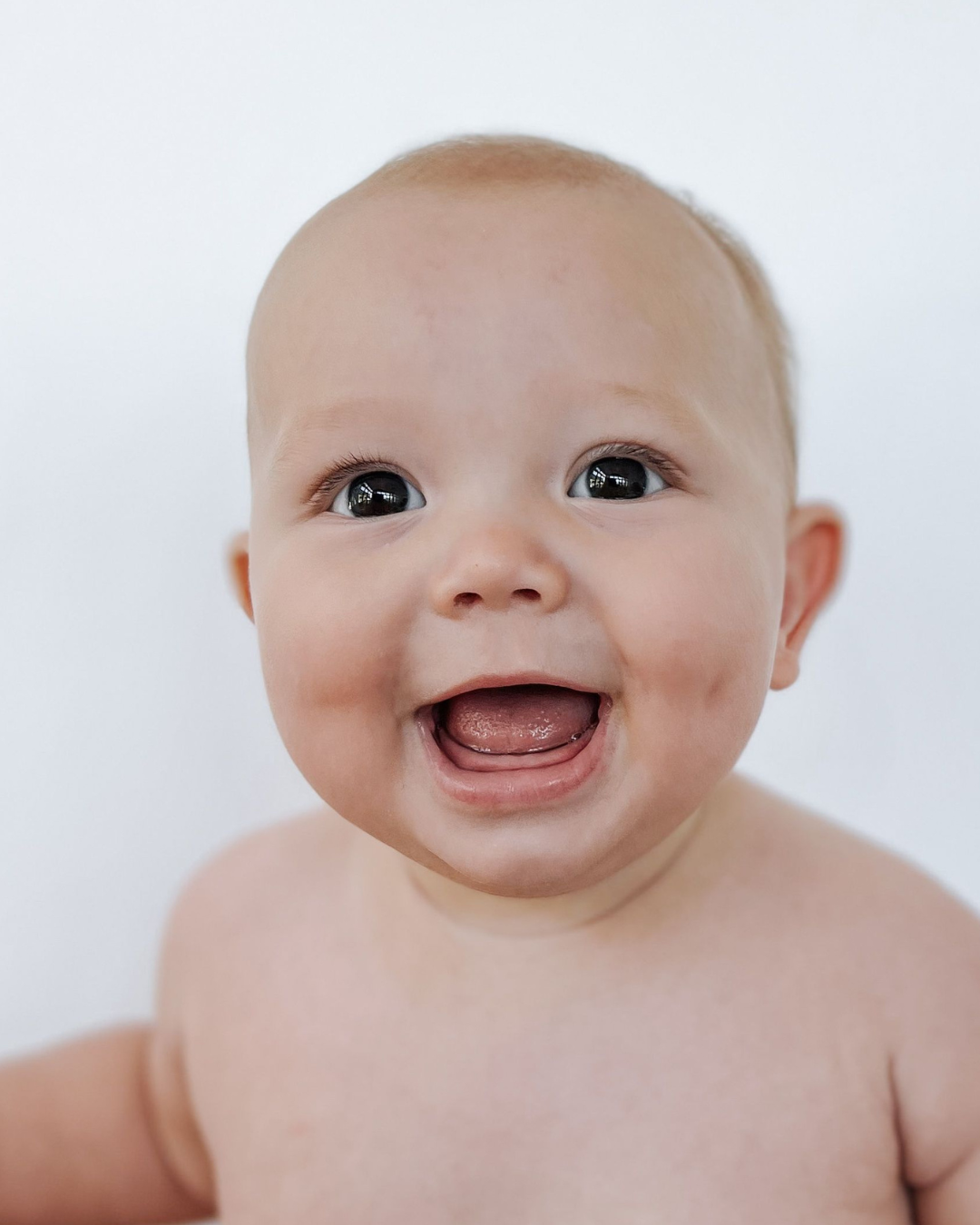 Close-up of a happy baby with big eyes and open mouth, showing a joyful expression against a plain white background.
