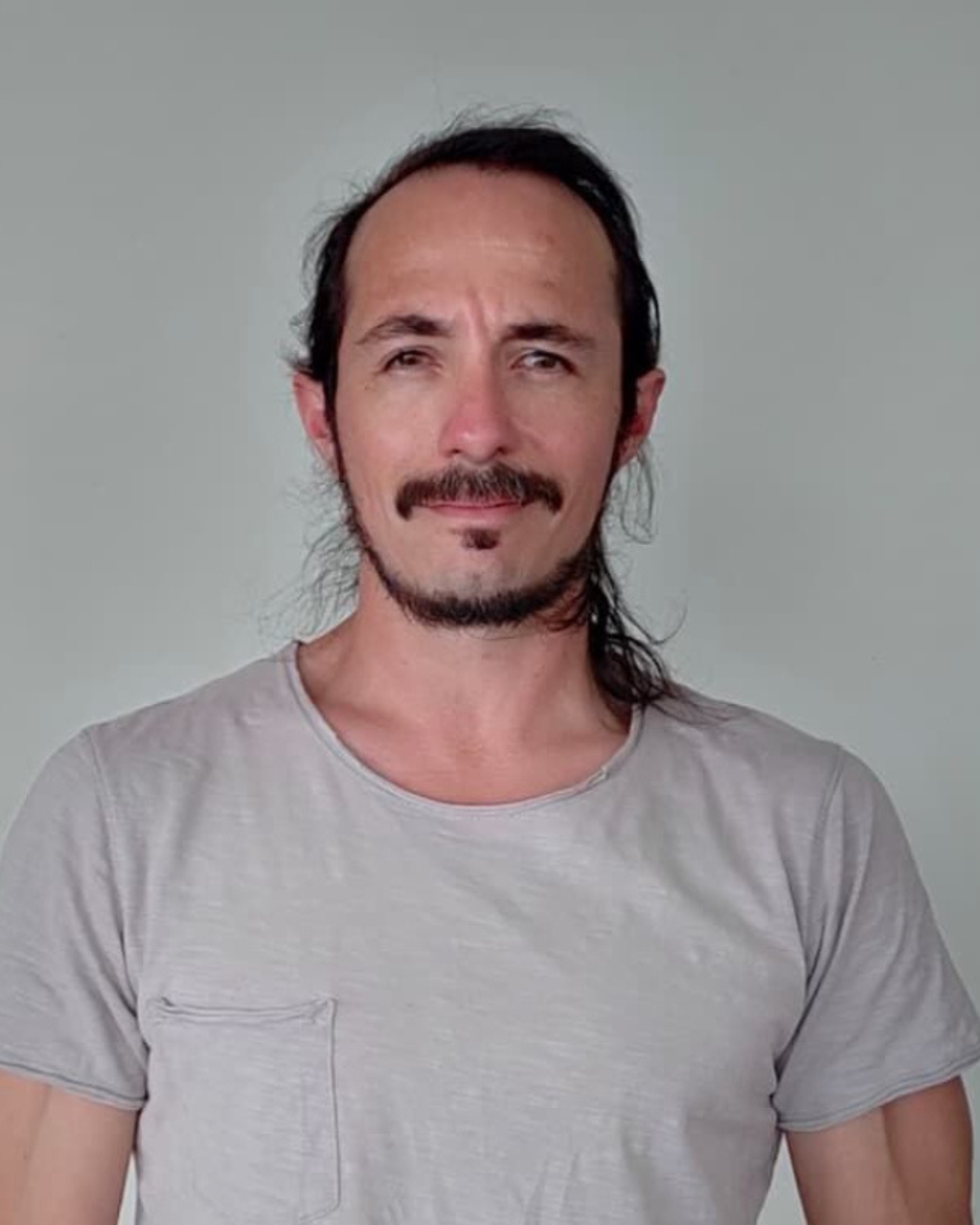 A man with long dark hair, a mustache, and goatee, wearing a light gray t-shirt, standing against a plain light gray background.
