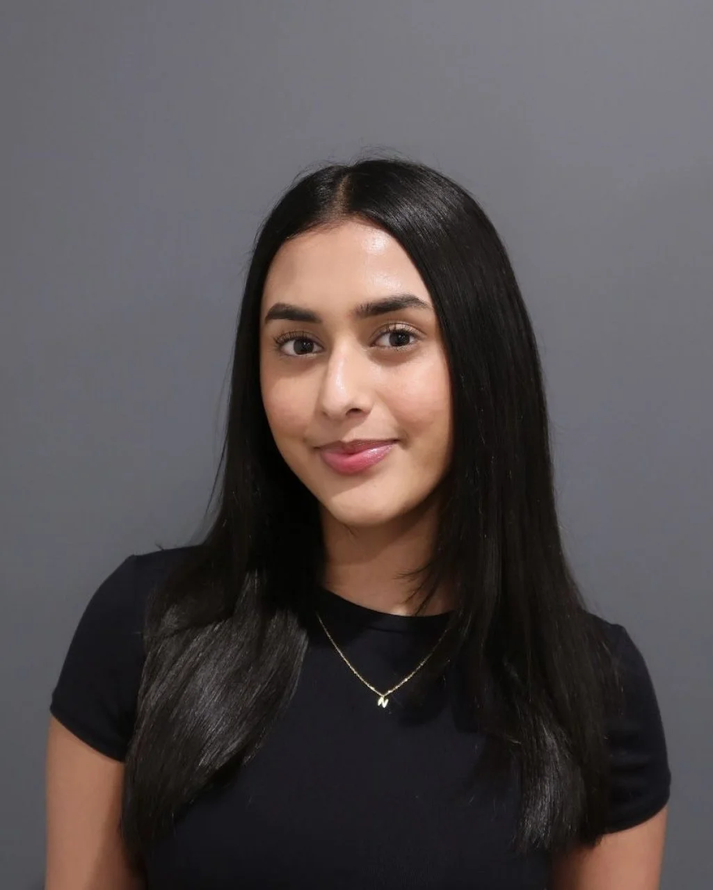 A young woman with long black hair, wearing a black top and a gold necklace, smiling and looking at the camera against a gray background.