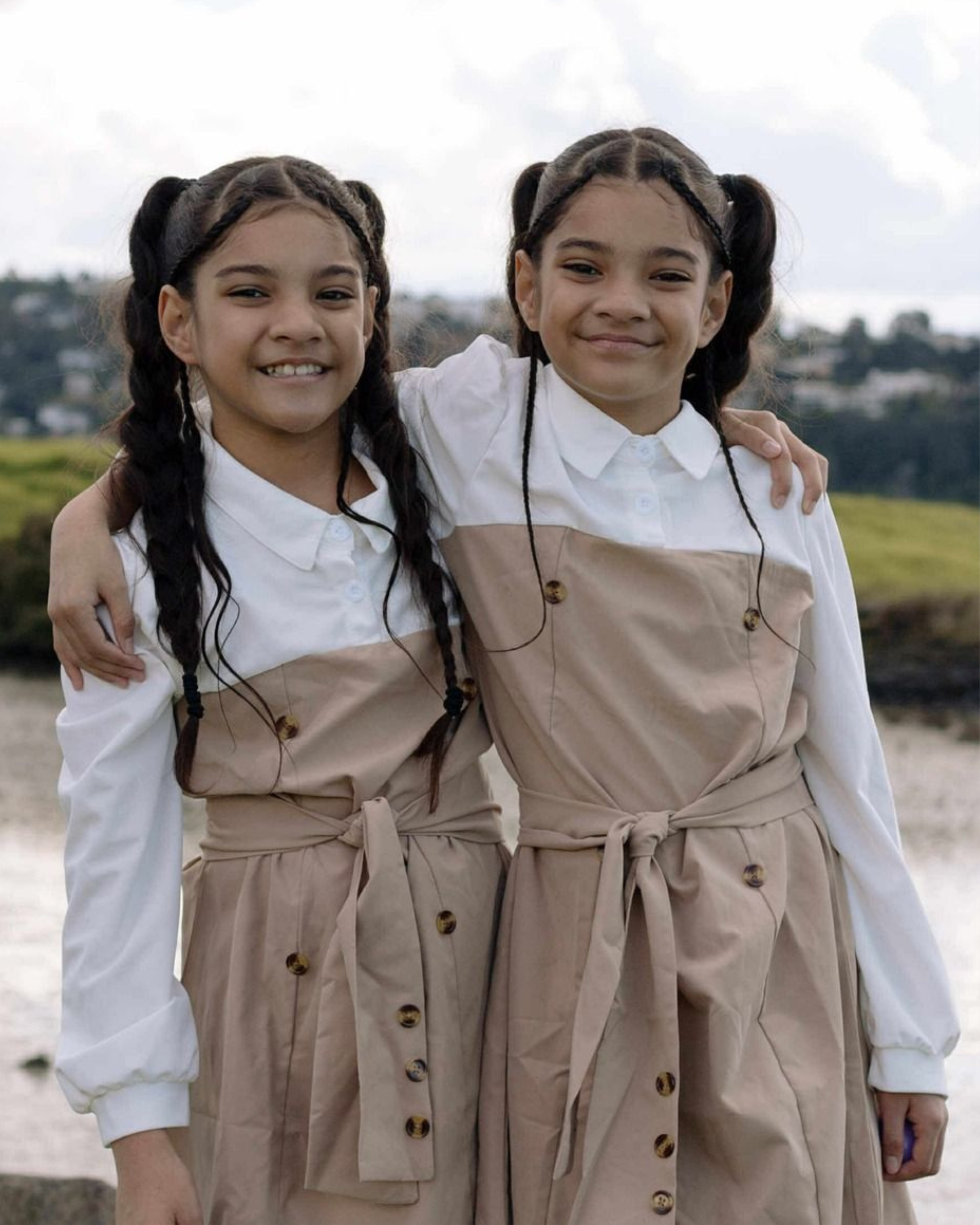 Two young girls with braided hair and light skin, wearing coordinated beige and white outfits, standing outdoors with arms around each other and smiling.