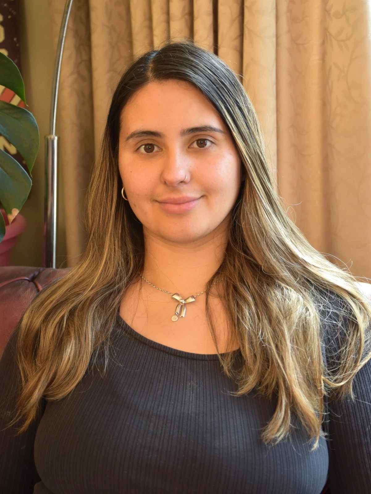 A young woman with long, wavy hair sitting indoors near a window with beige curtains, wearing a black top and a necklace.