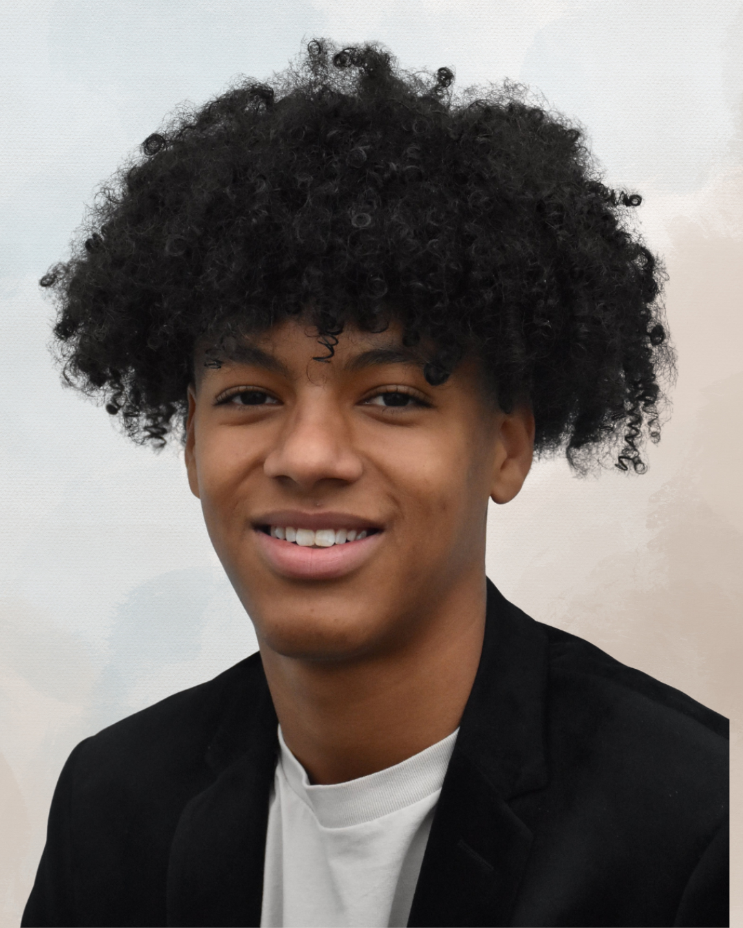 Portrait of a young man with curly black hair, smiling, wearing a black blazer and white shirt, against a neutral background.