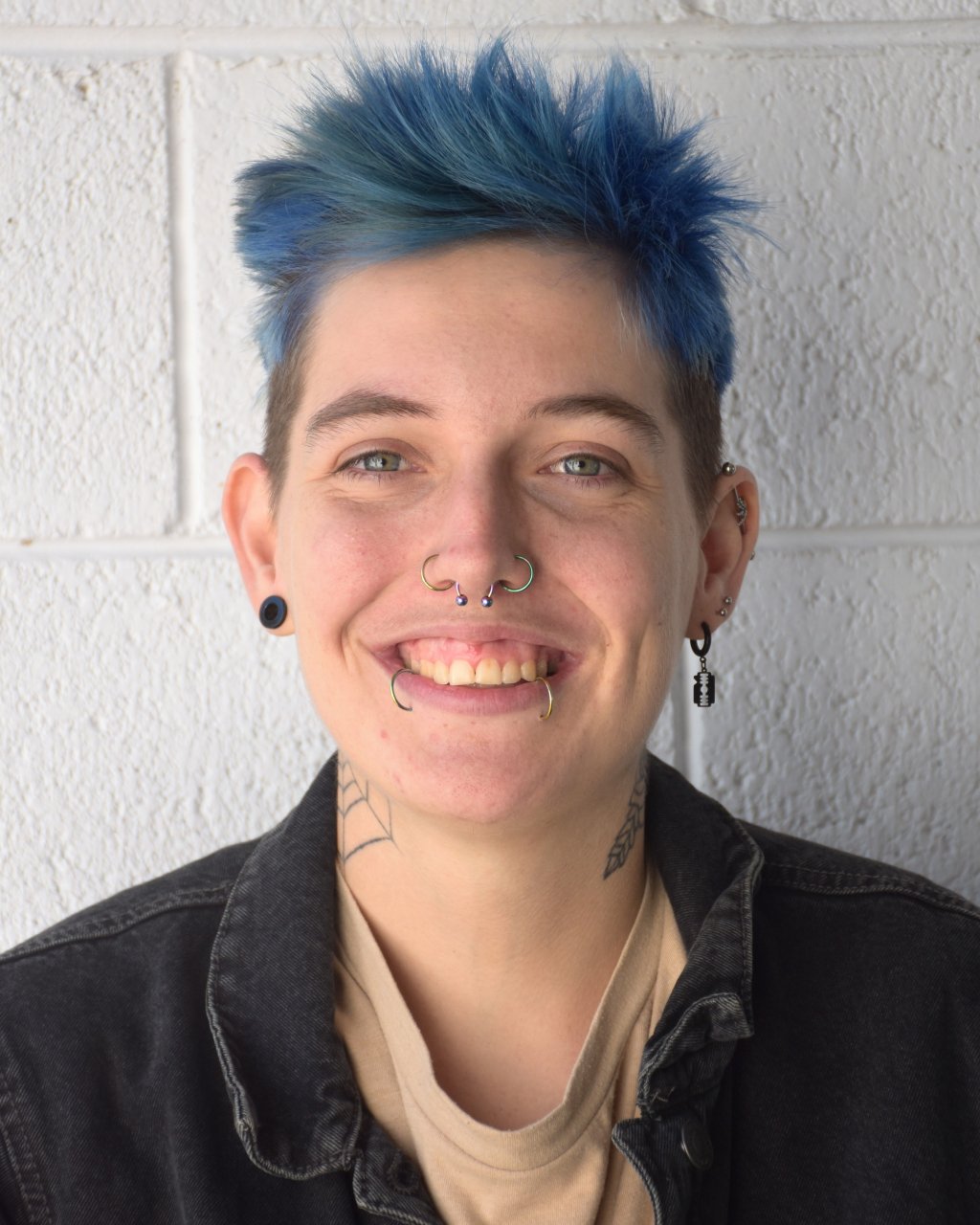 A young person with short brown hair, blue eyes, freckles, and earrings, wearing a black shirt, standing against a plain light-colored wall.