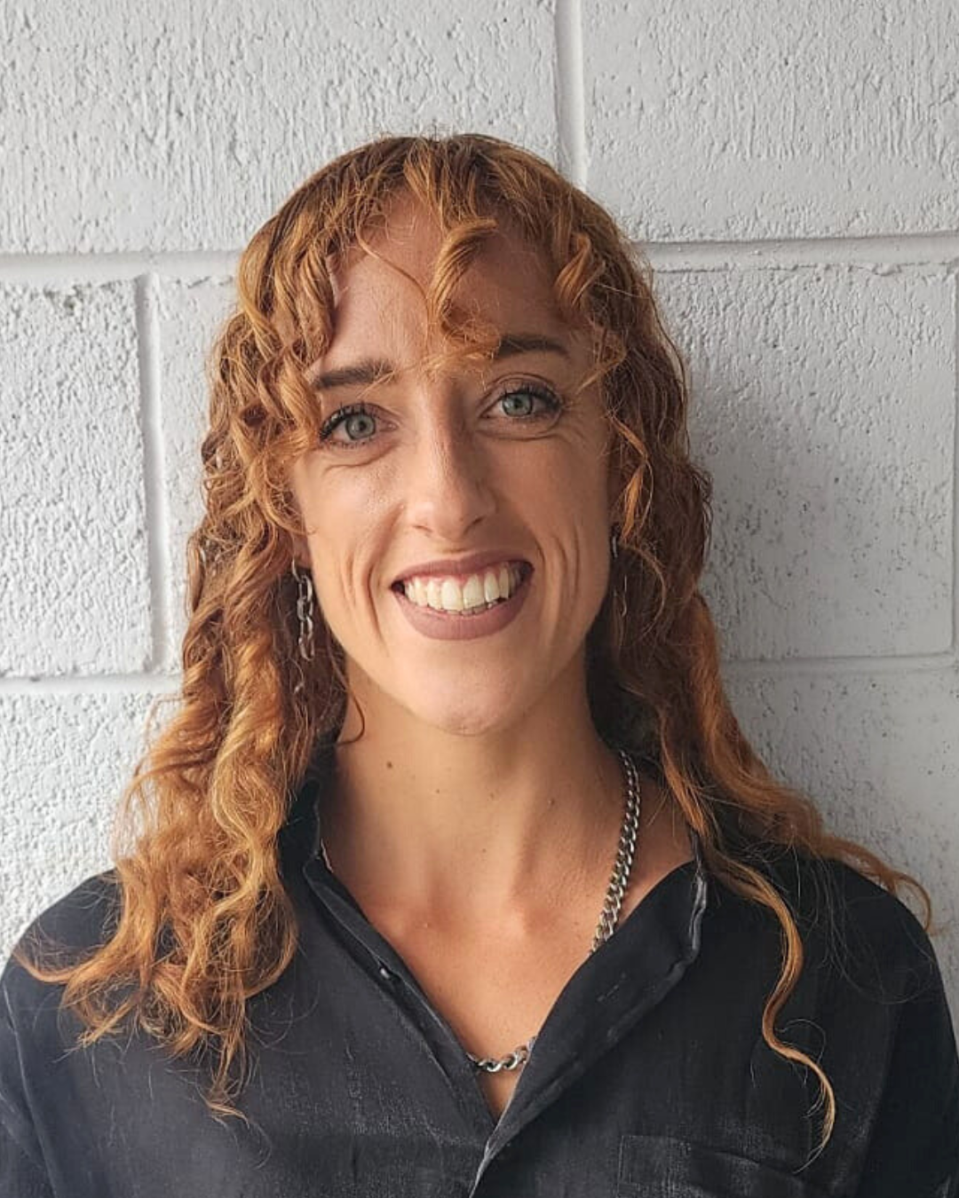 A woman with curly red hair wearing a black, beige, and teal sequin top, standing in front of a wall decorated with colorful artwork.
