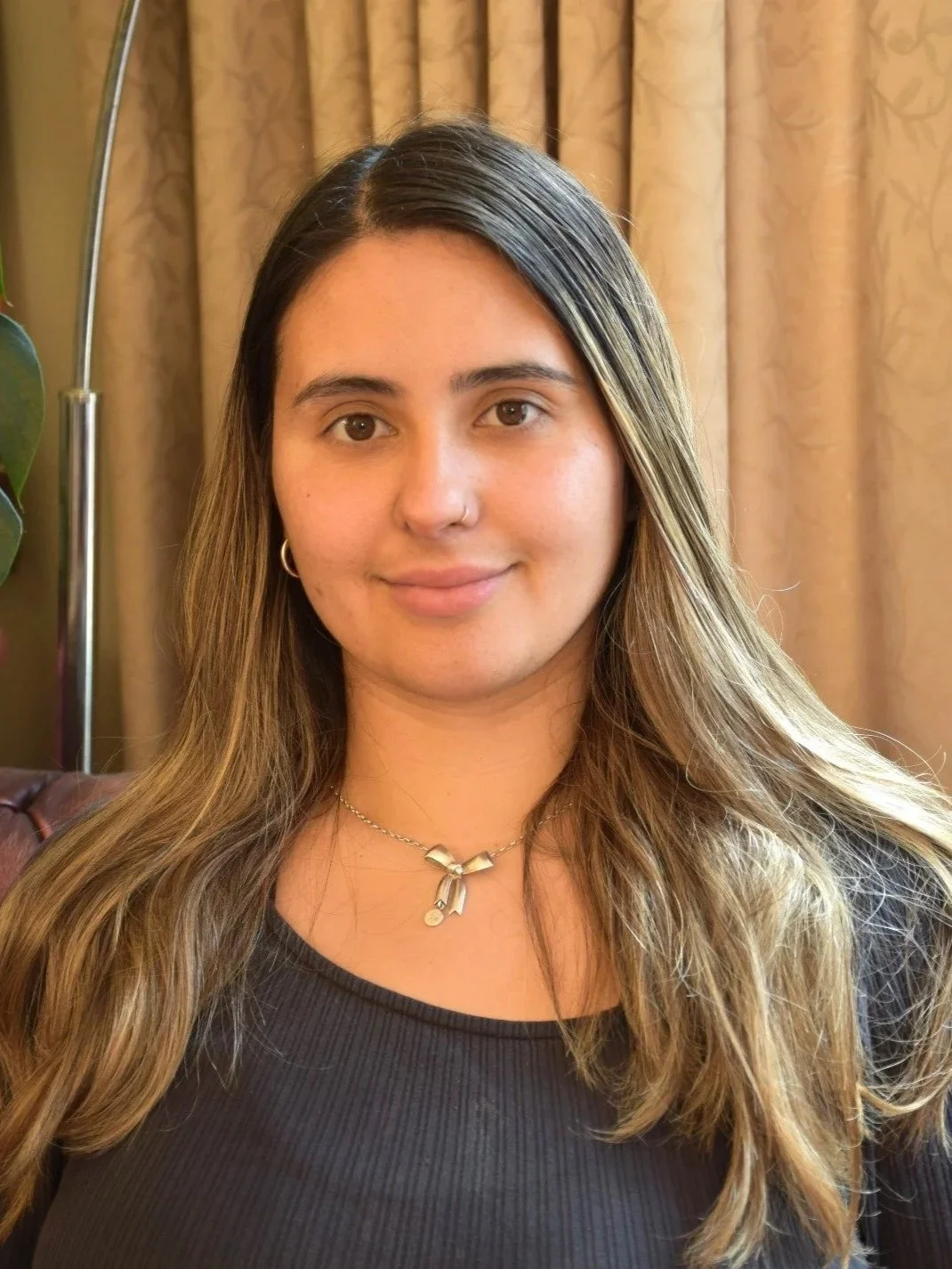 A young woman with long brown hair and a nose piercing, wearing a black top and a silver necklace, smiling indoors with beige curtains in the background.