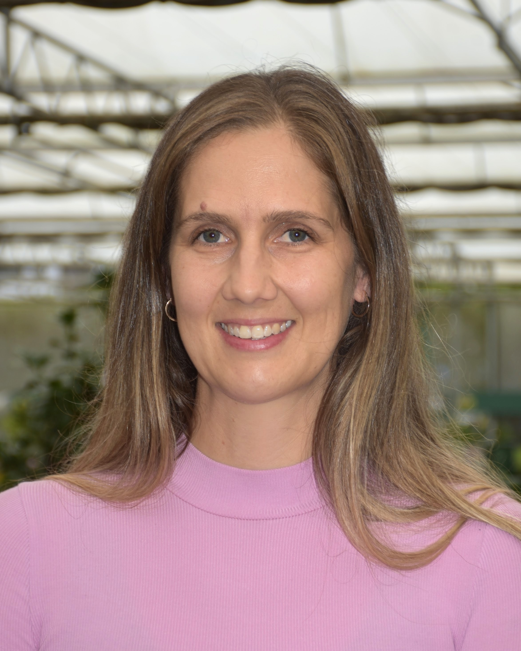 A woman with light brown hair and blue eyes wearing a pink top, smiling in a greenhouse with plants and a glass ceiling in the background.